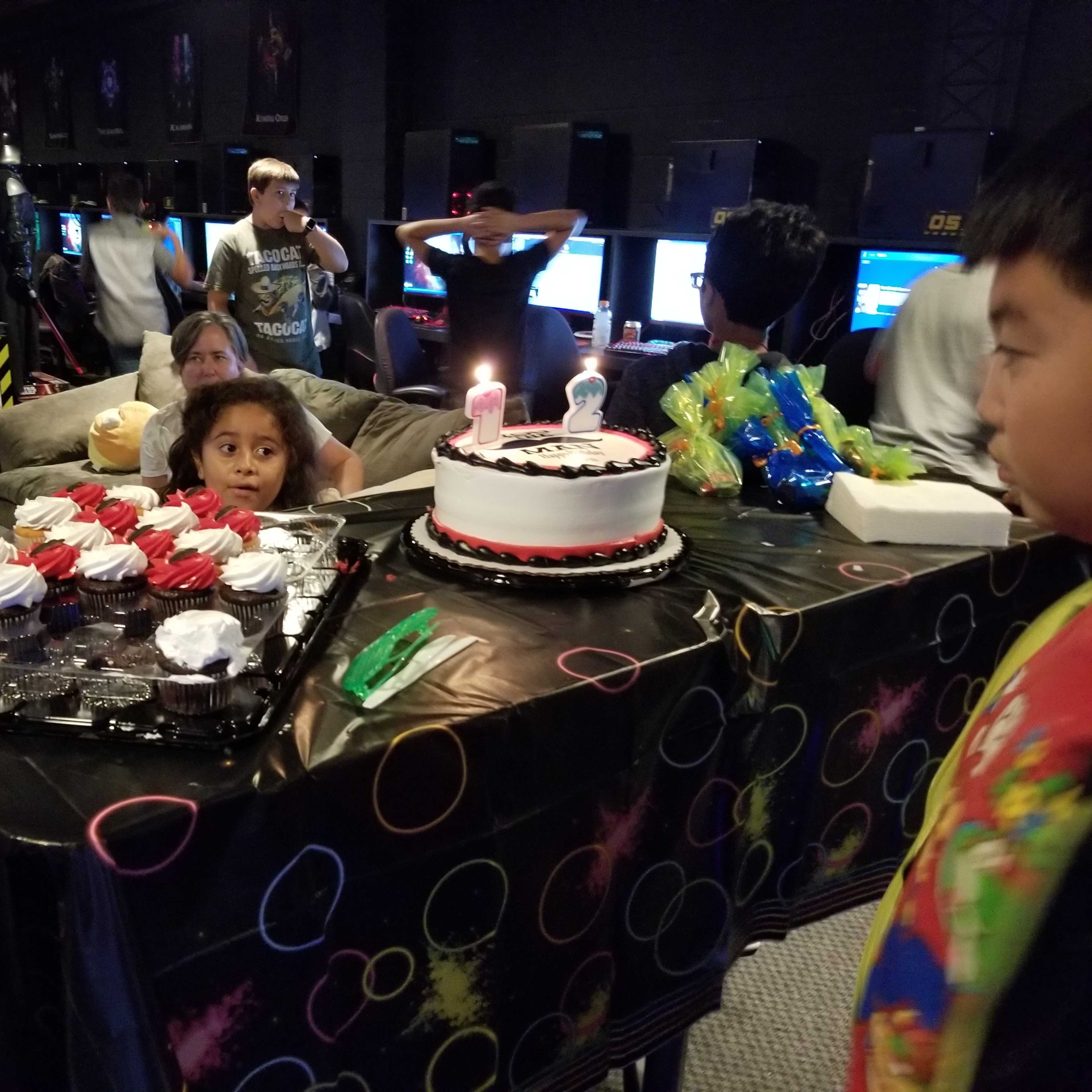 A birthday celebration takes place in a gaming lounge with a table set up featuring a decorated cake and cupcakes, surrounded by children and gaming monitors in the background.