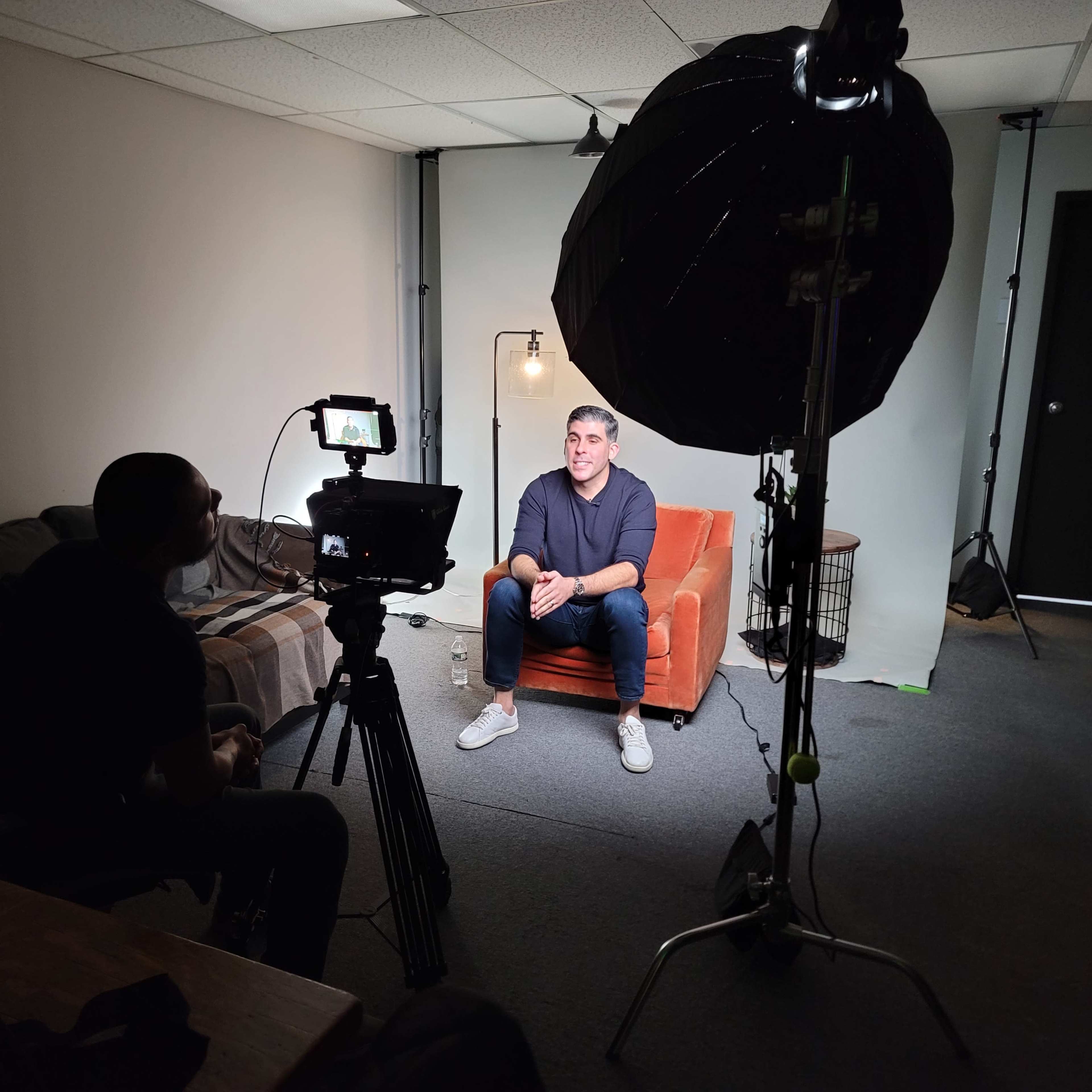 A person is being filmed while sitting on an orange couch in a dimly lit room, with a camera and lighting equipment set up in front of them.