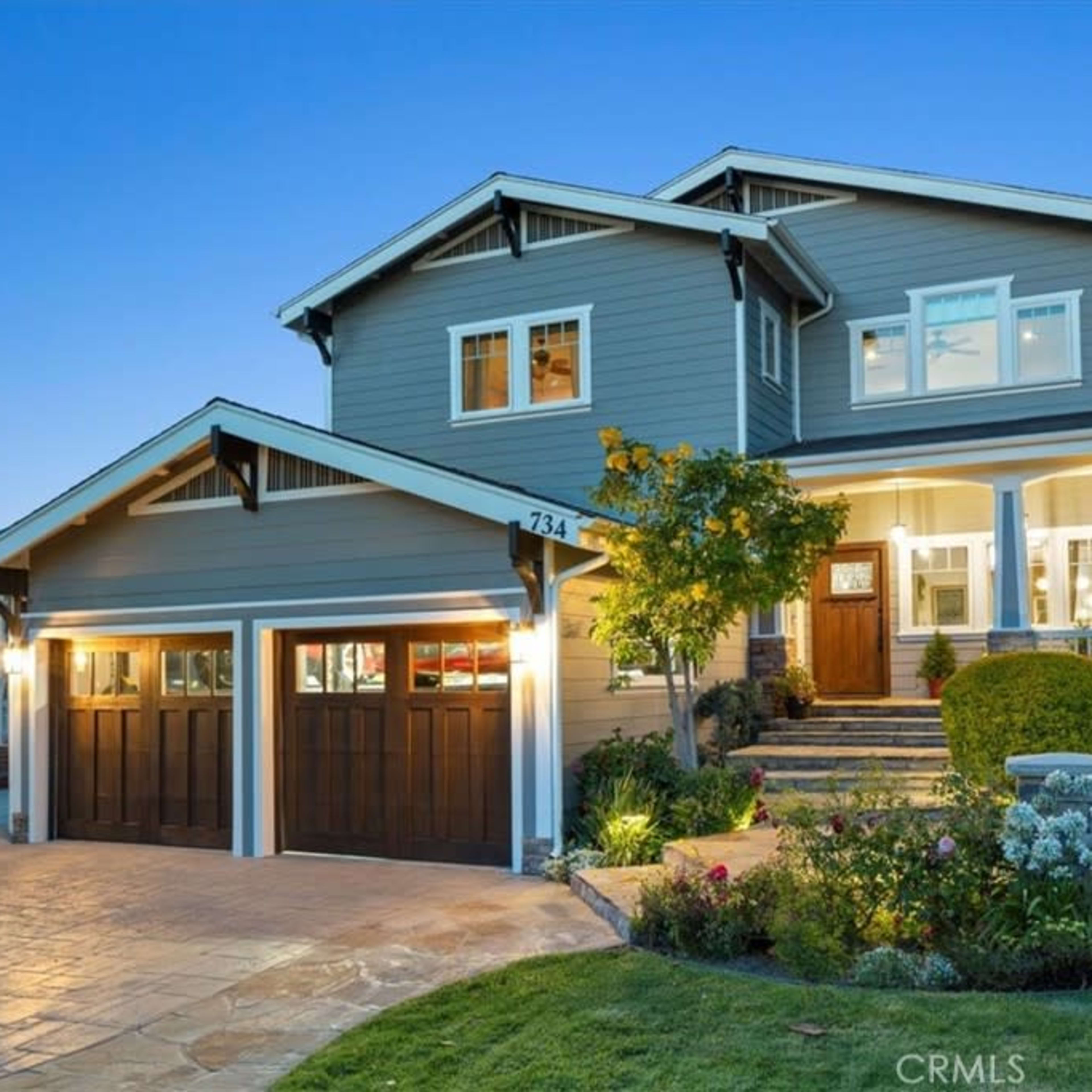 A two-story house with a front porch and manicured landscaping is illuminated at dusk.