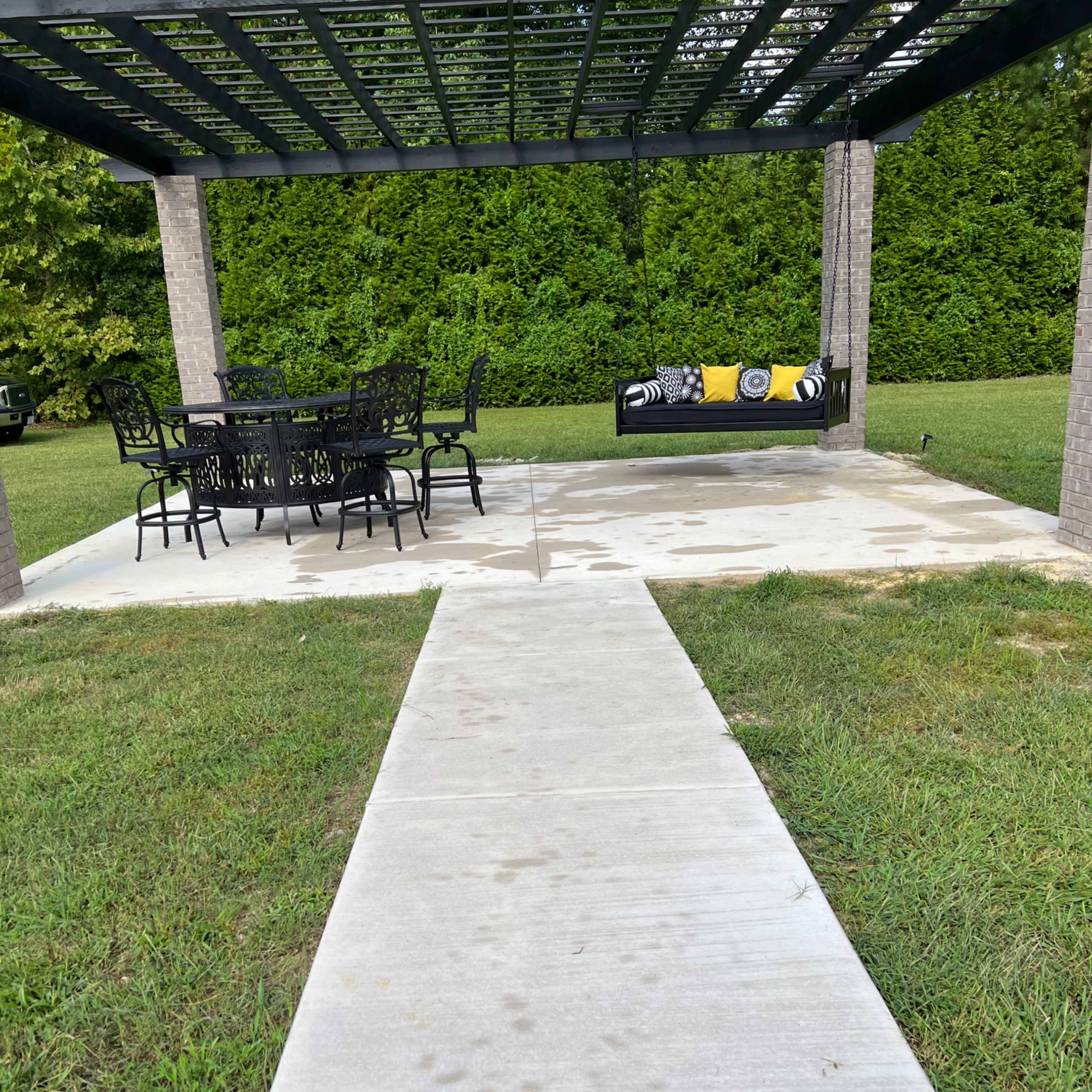 A concrete pathway leads to a covered patio featuring a dining table and a hanging bench surrounded by grass.