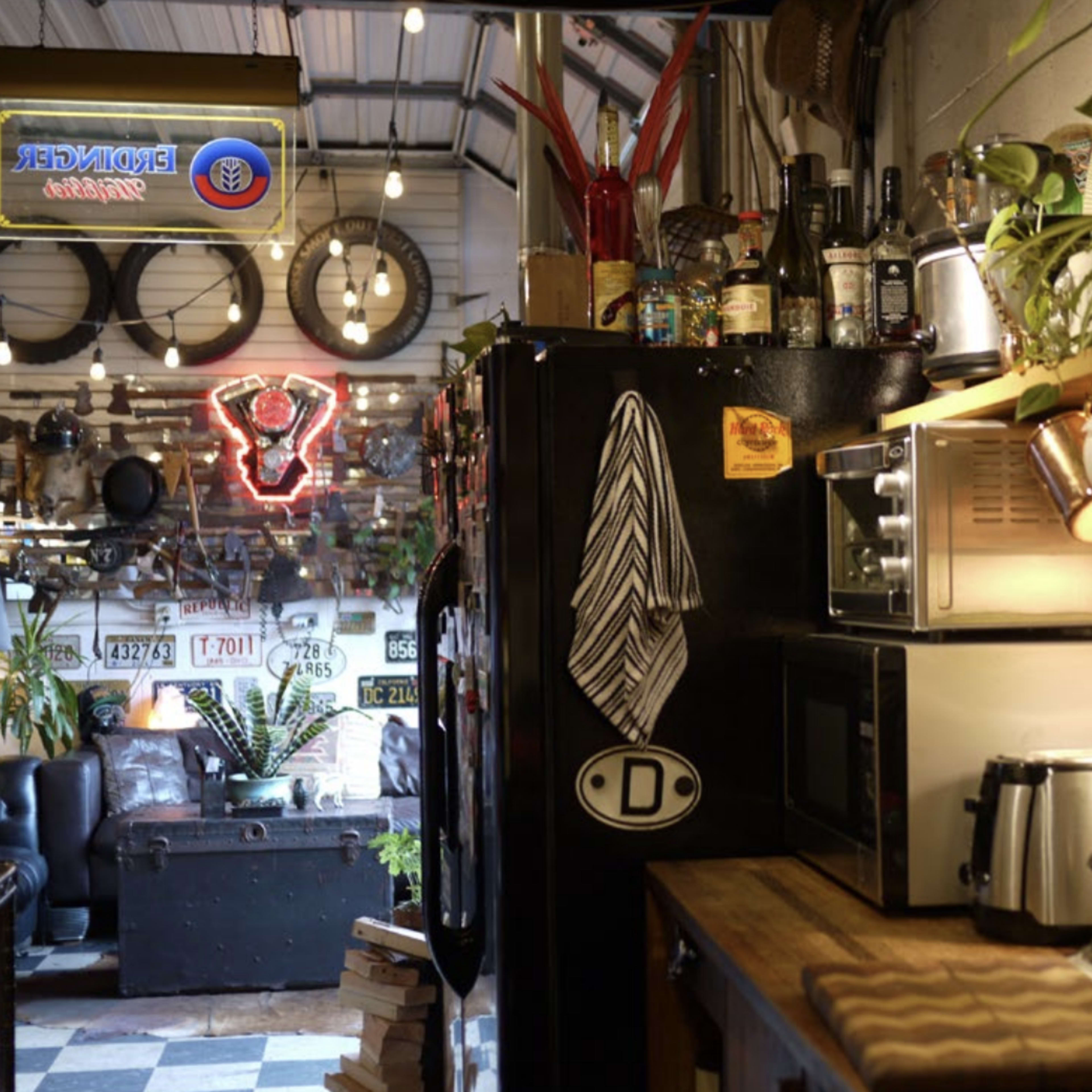 A rustic kitchen area with a black refrigerator, wooden shelves, various kitchen appliances, and plants, all illuminated by warm lights and neon signs.