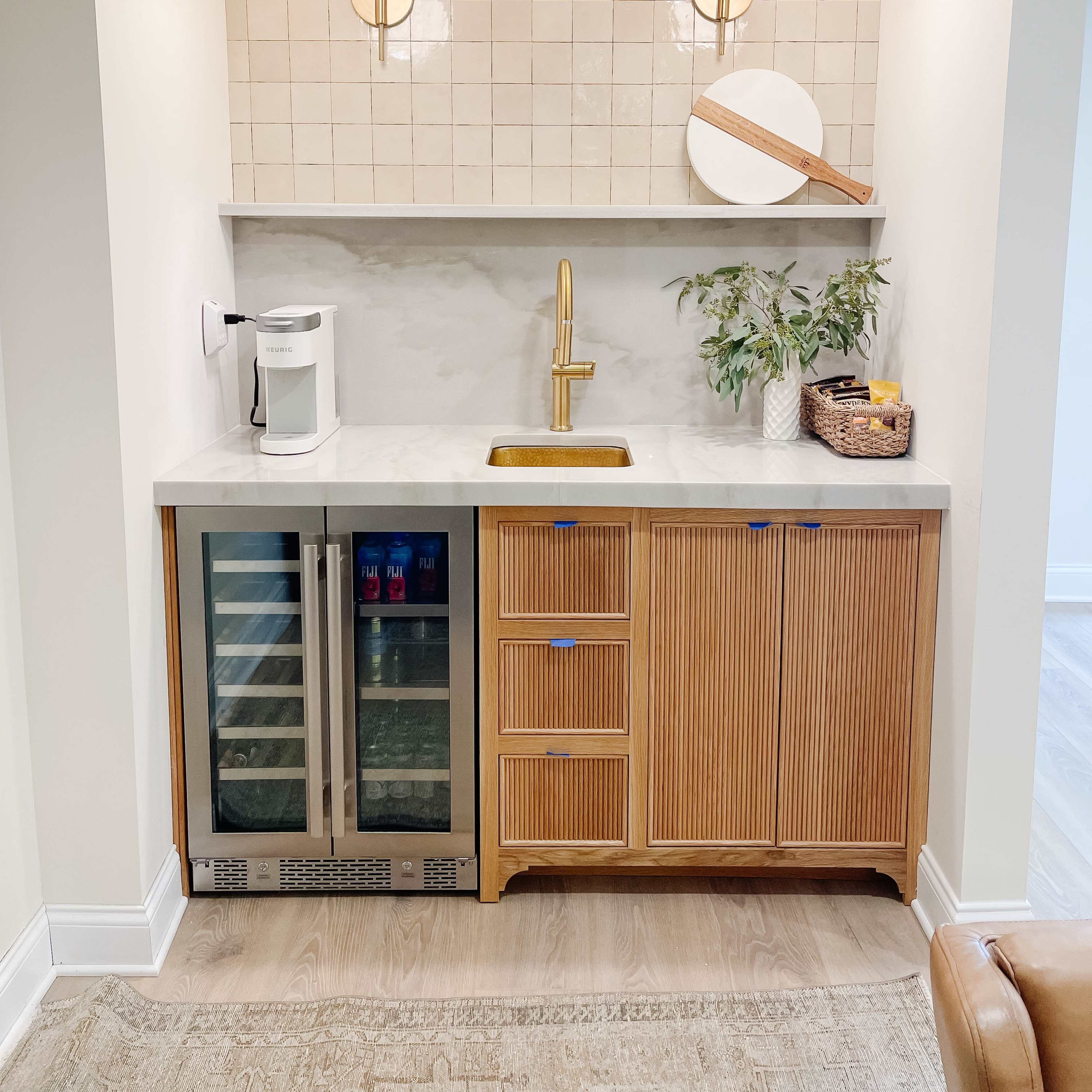 A modern kitchenette featuring a countertop sink, a mini fridge, wooden cabinetry with vertical slats, and decorative light fixtures above.