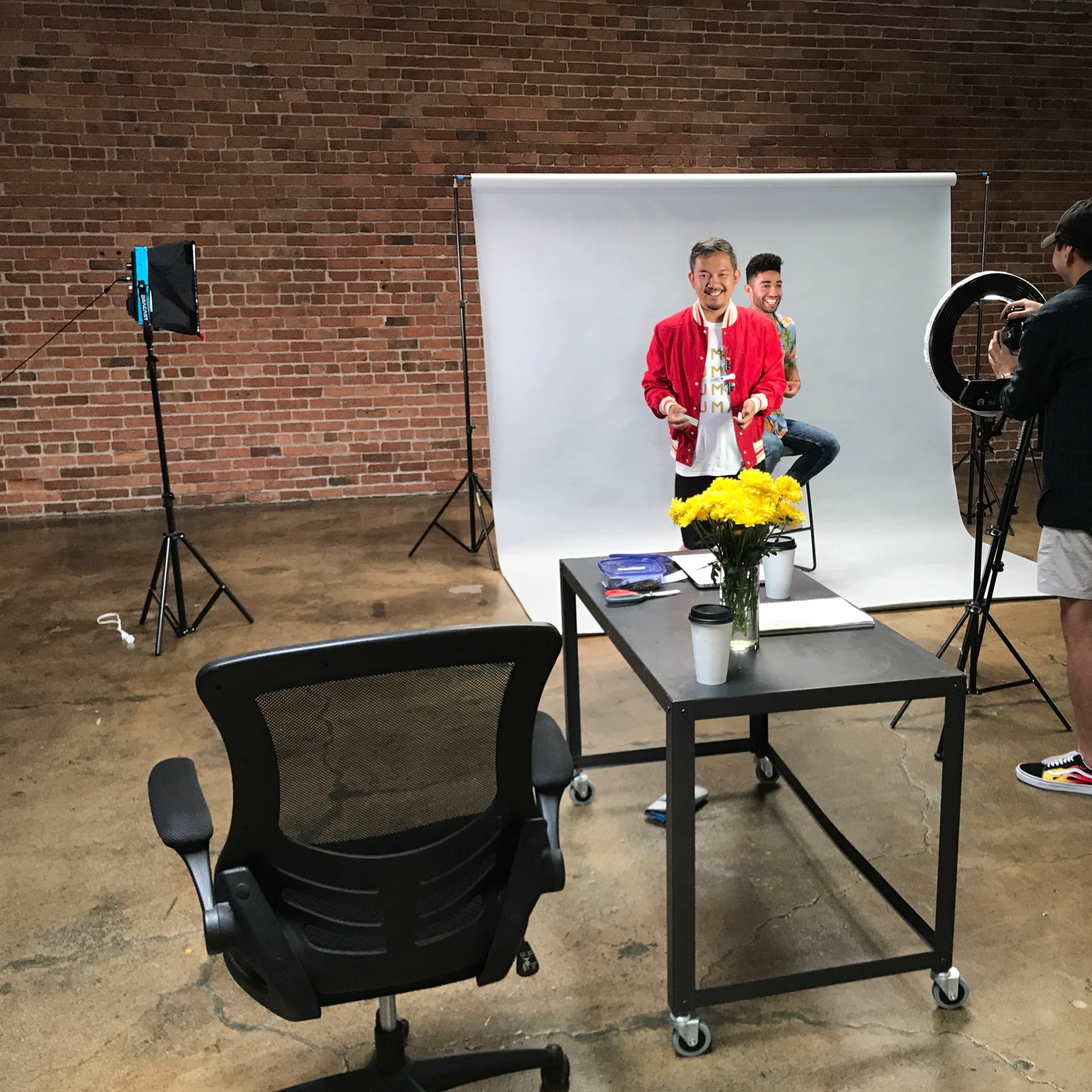 A photo shoot is taking place in a studio with a backdrop, featuring a man in a red jacket and another person in the background, while a third person adjusts lighting equipment.