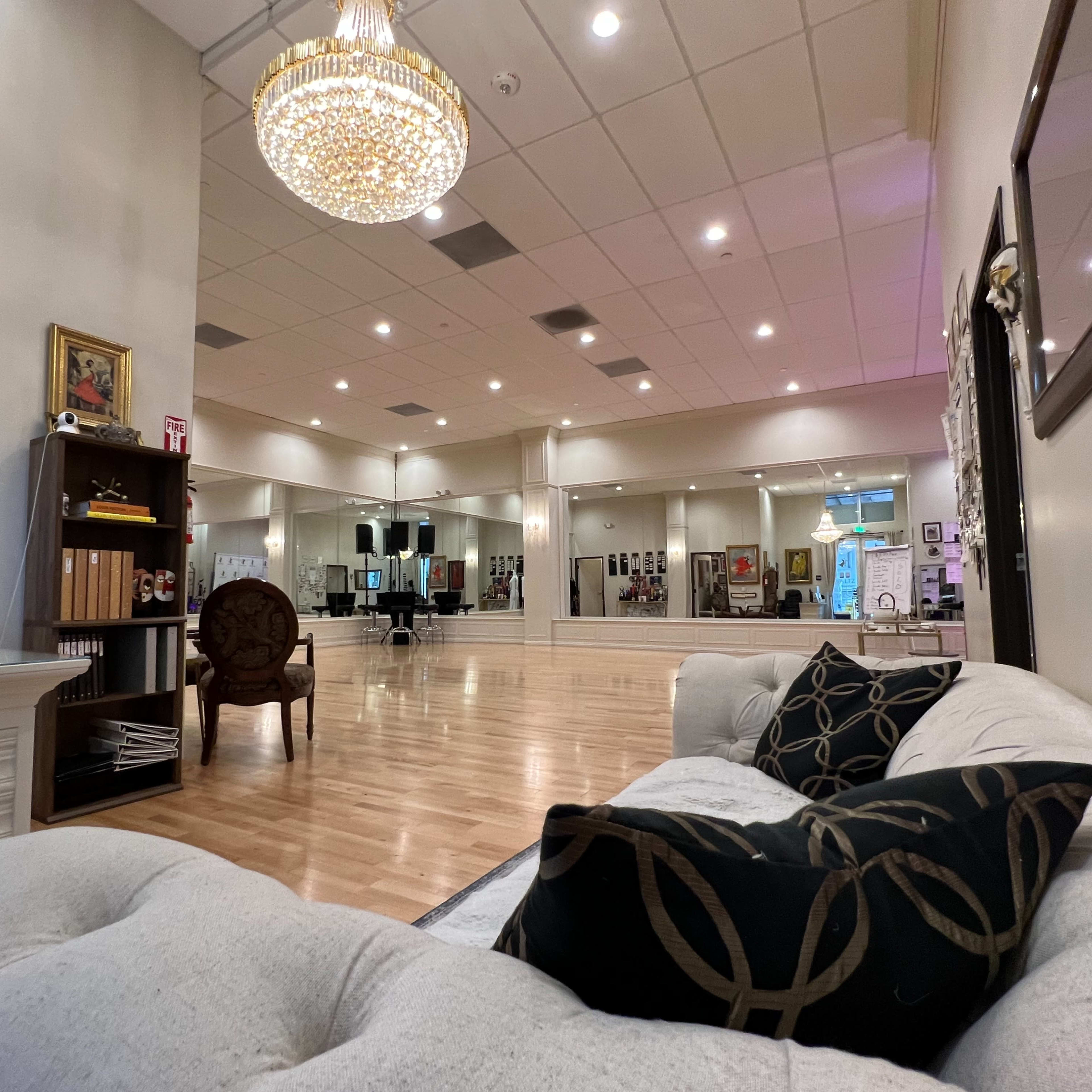 A spacious dance studio with wooden flooring, mirrors on the walls, and a chandelier overhead, featuring a comfortable couch and a decorative chair in the foreground.