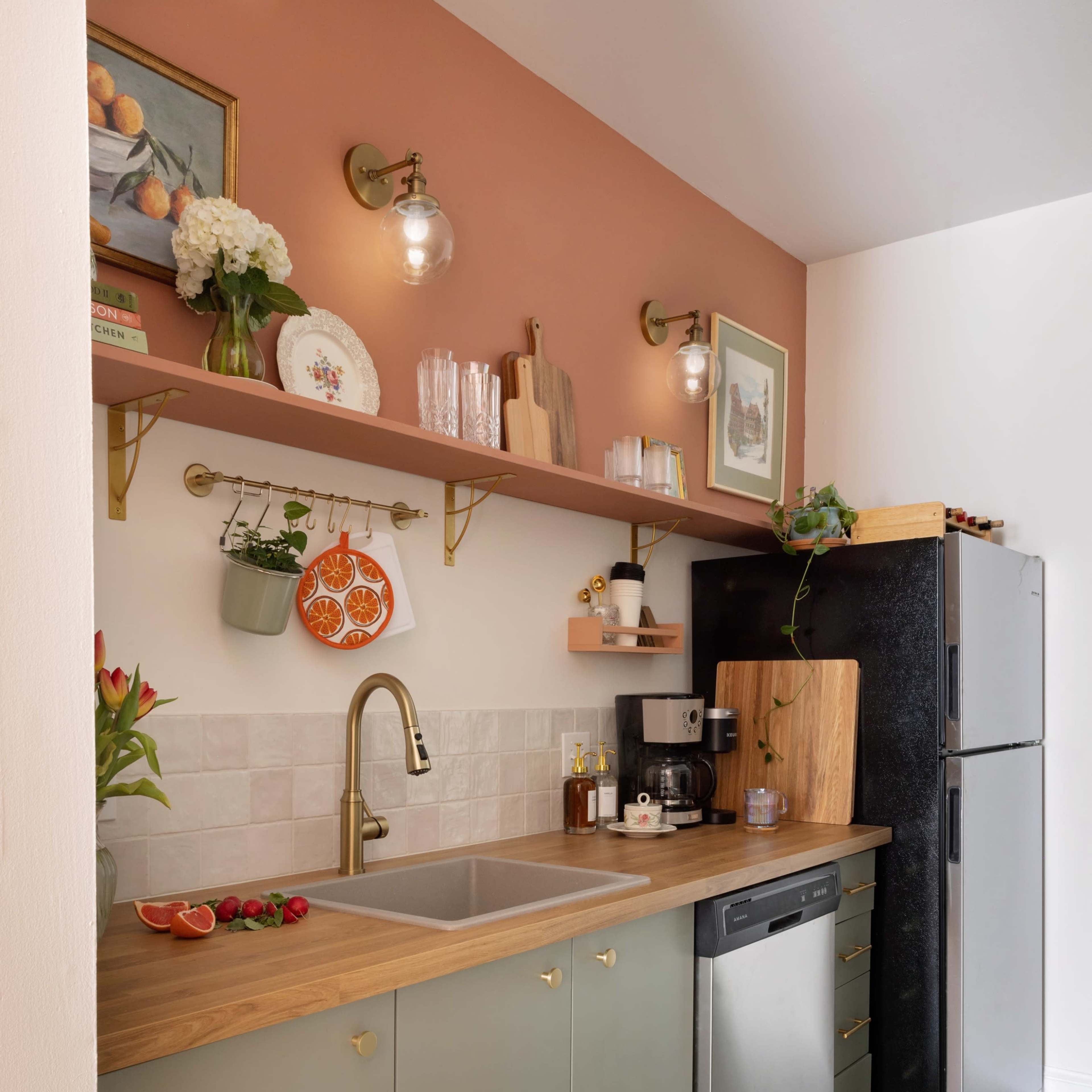 A modern kitchen featuring a double sink with a gold faucet, a coffee maker, a refrigerator, and open shelving displaying kitchenware against a peach-colored wall.