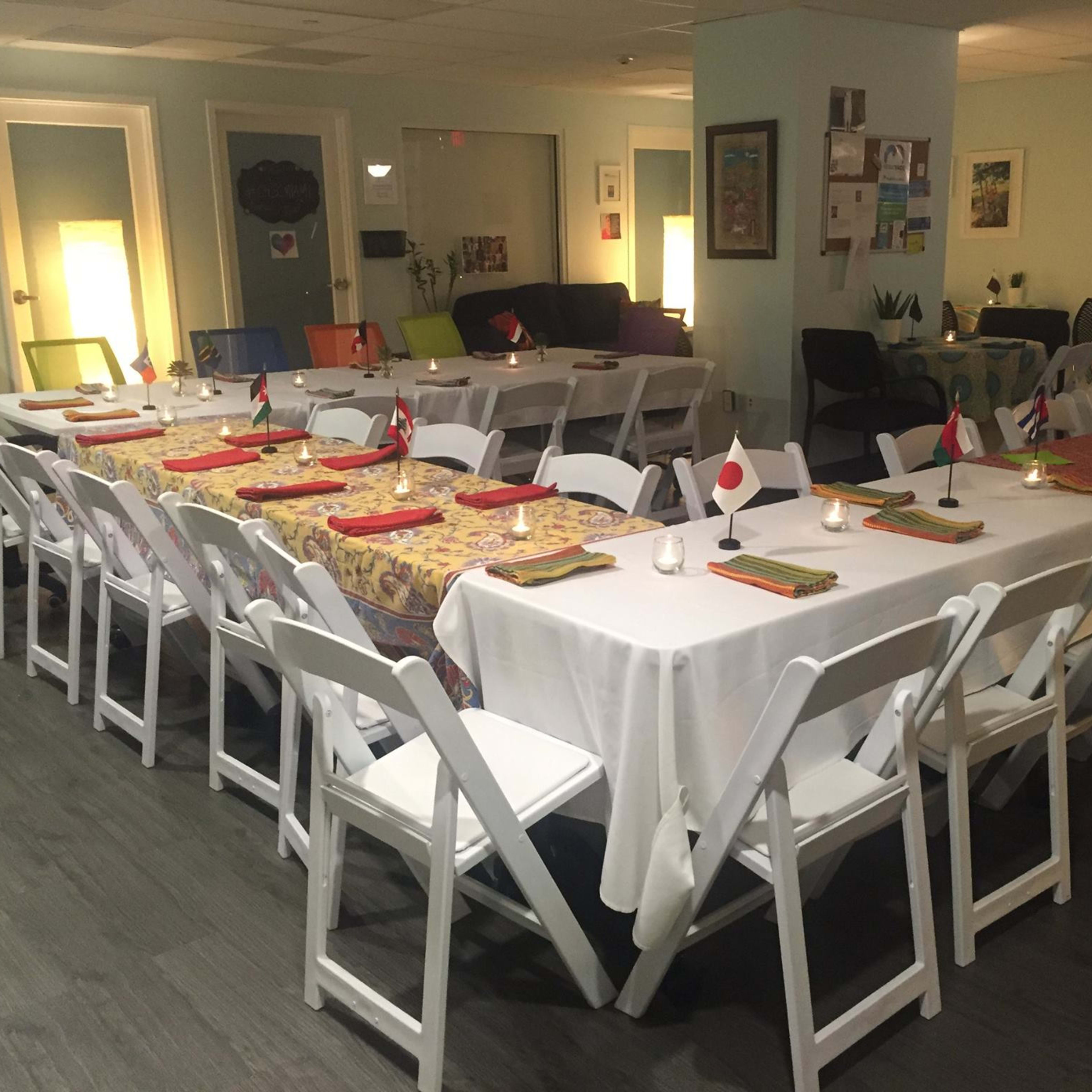 A dining area is set up with multiple tables arranged in a U-shape, adorned with tableware and flags from various countries, ready for a gathering.
