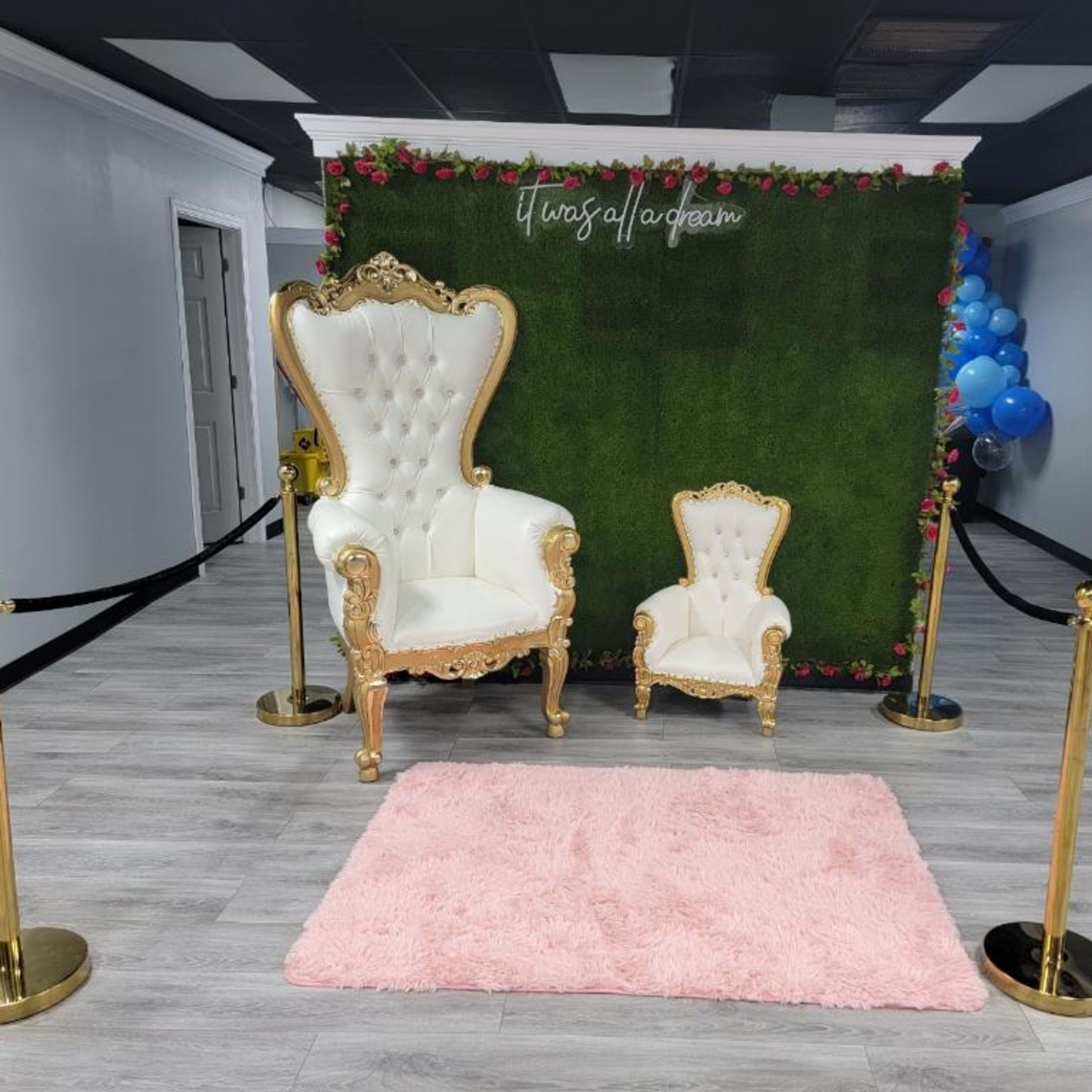 A decorated area featuring a large ornate white chair and a smaller matching chair in front of a green wall with roses, alongside a pink rug and stanchions.
