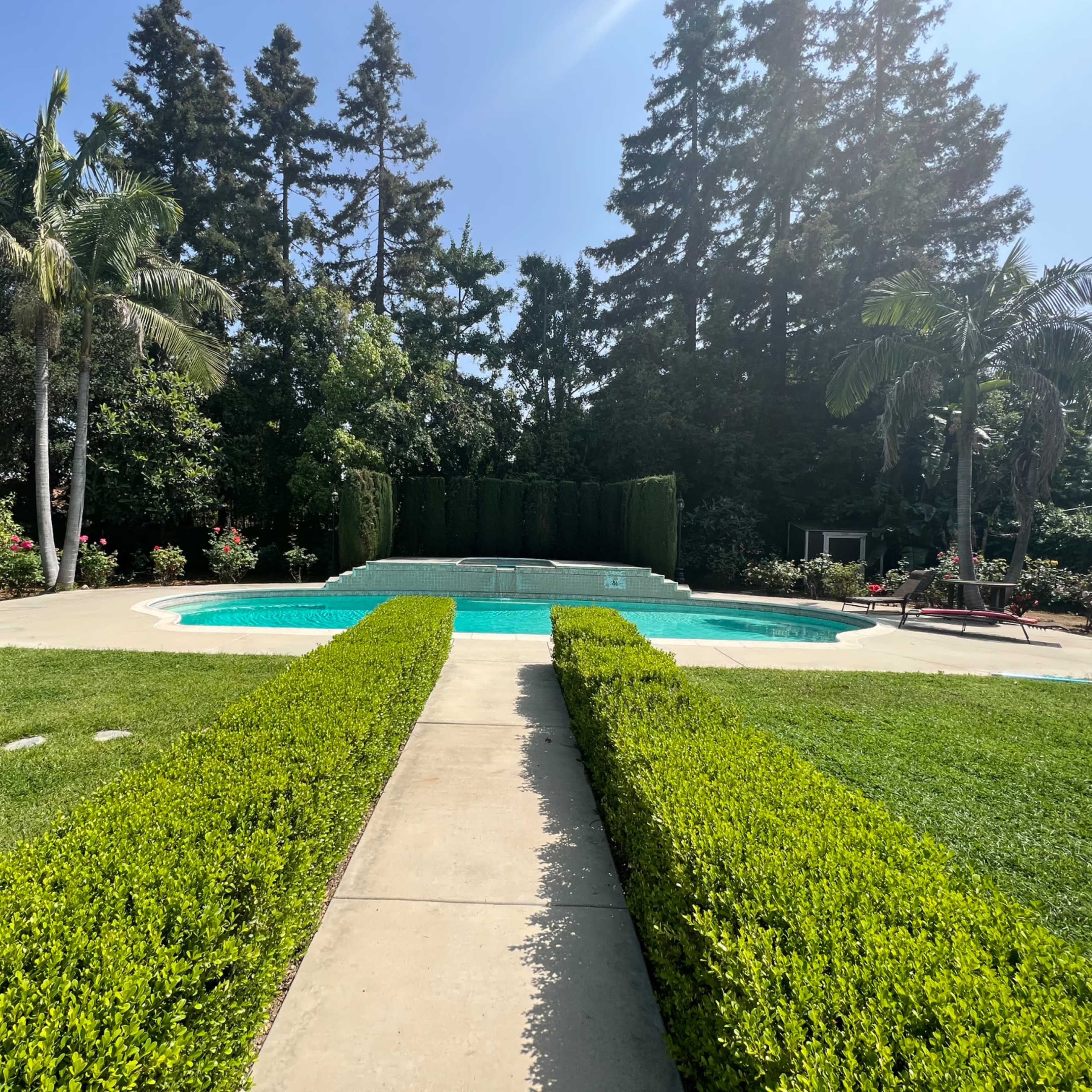 A pool is surrounded by neatly manicured hedges and a lawn, with tall trees and landscaped flower beds in the background.