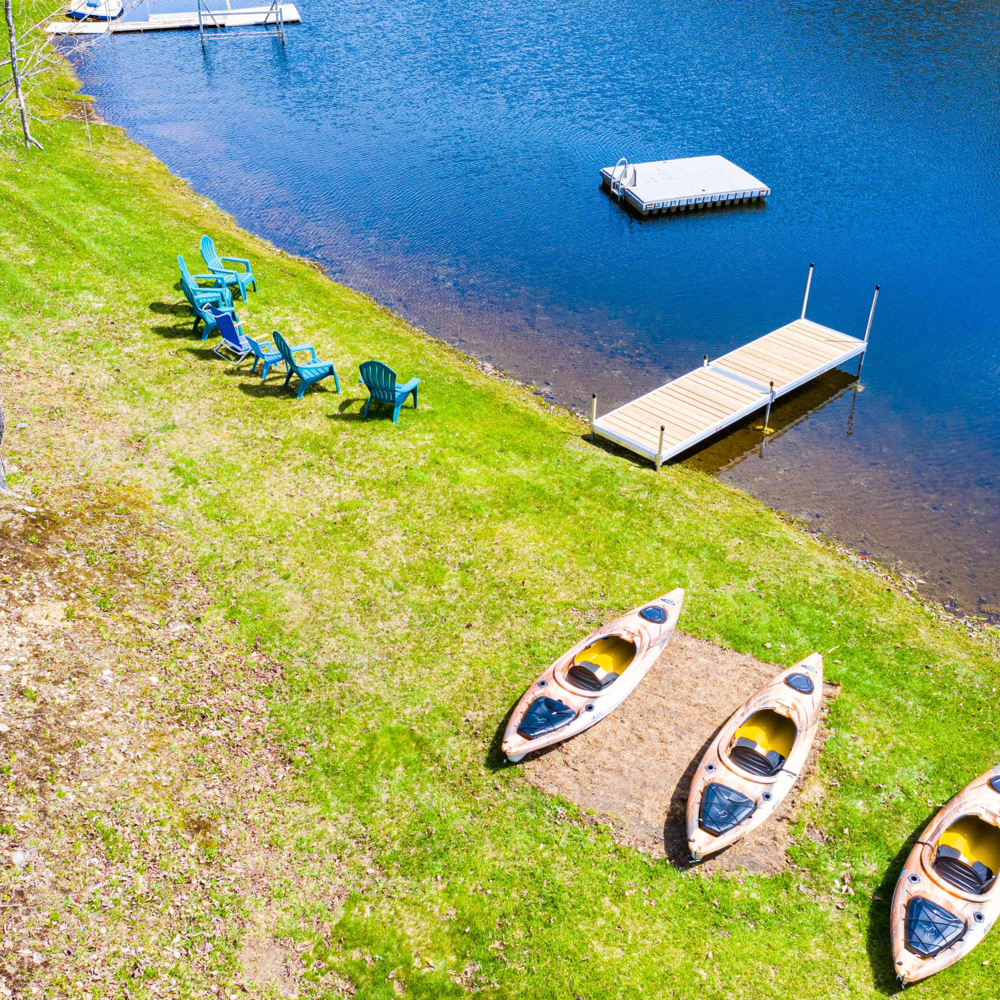 Three kayaks are positioned on the grass near a calm lake, alongside a set of blue Adirondack chairs and a wooden dock.