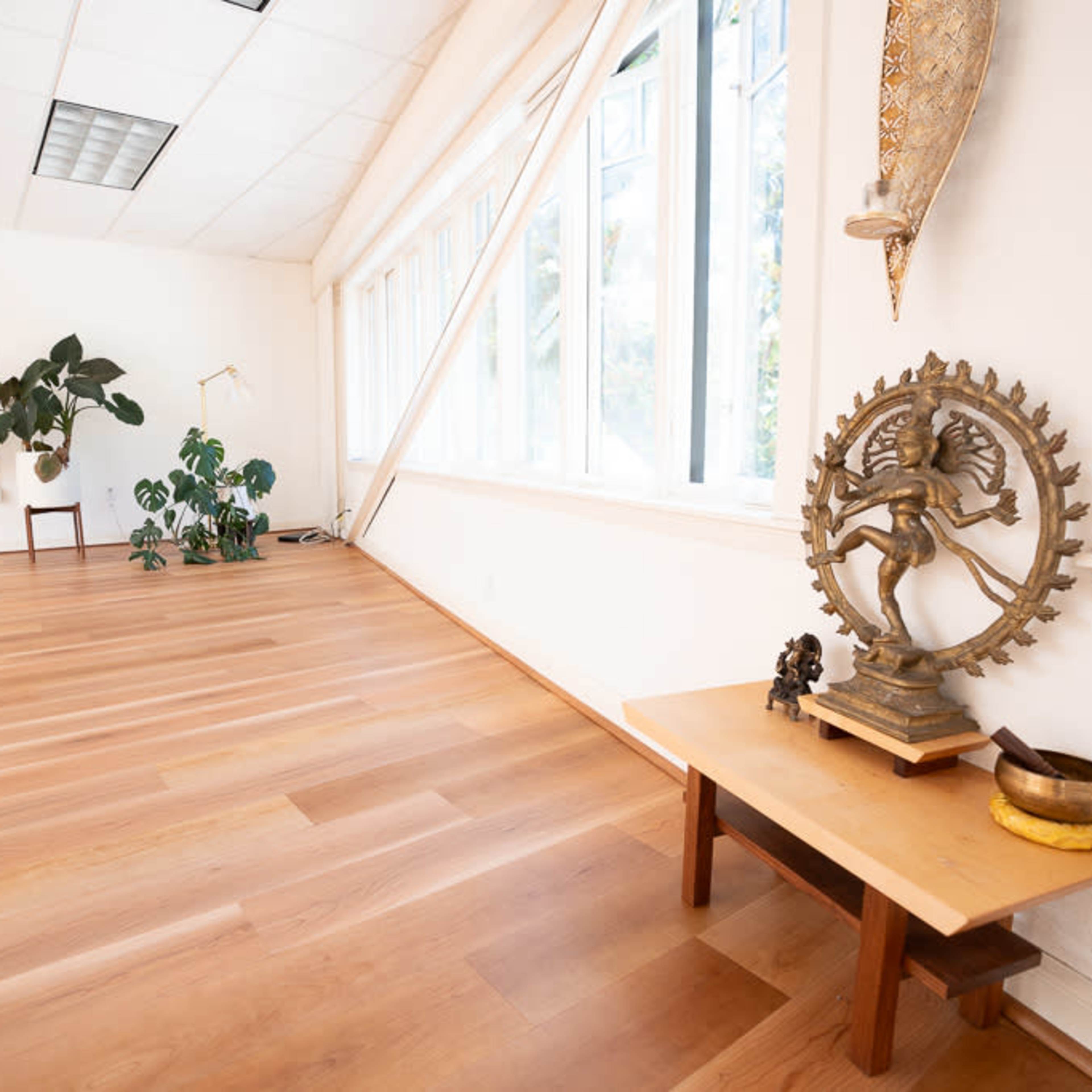 A spacious room with wooden flooring, large windows, and a decorative table featuring a brass statue and a bowl.