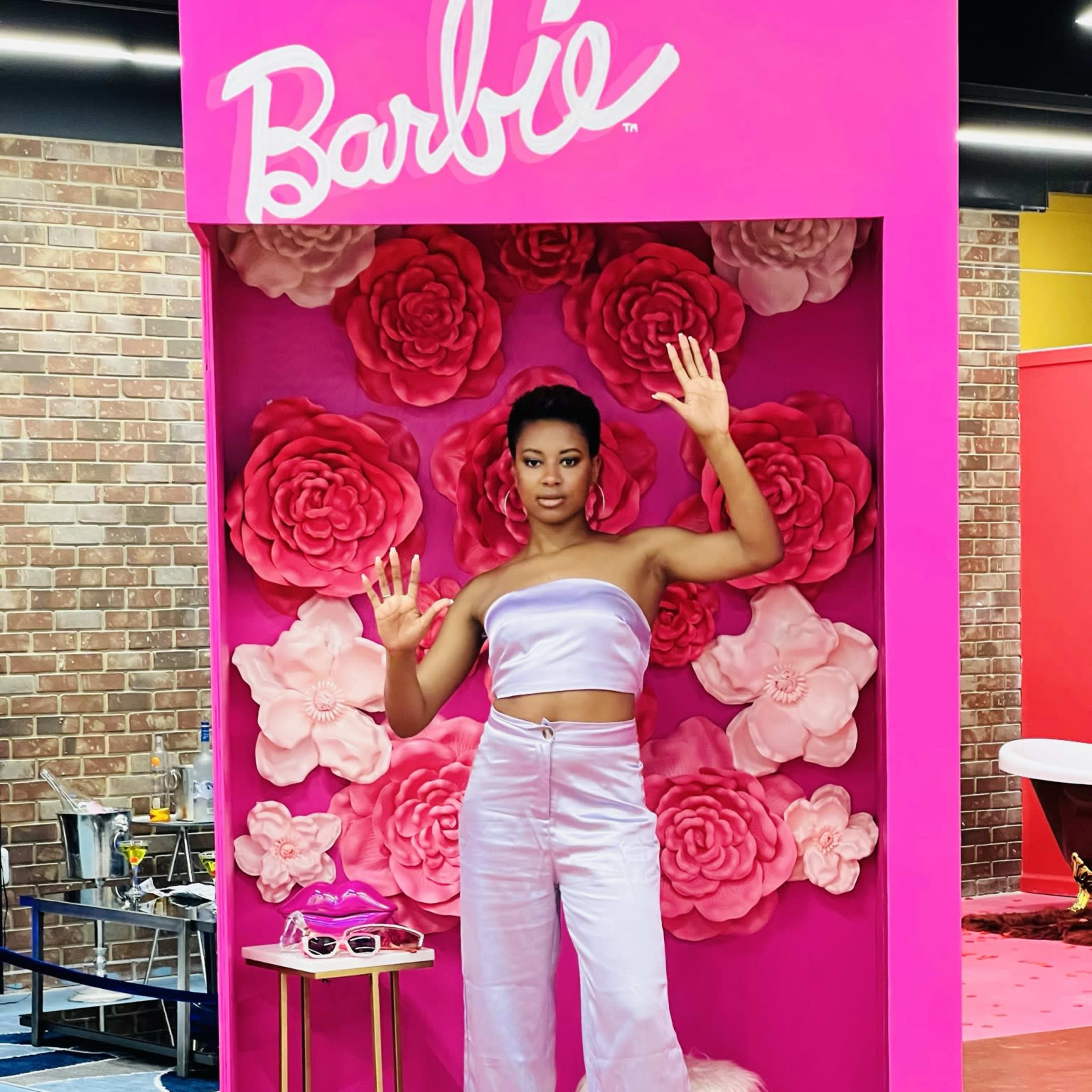 A model poses inside a pink display featuring large paper flowers and fashion accessories, wearing a lavender two-piece outfit.