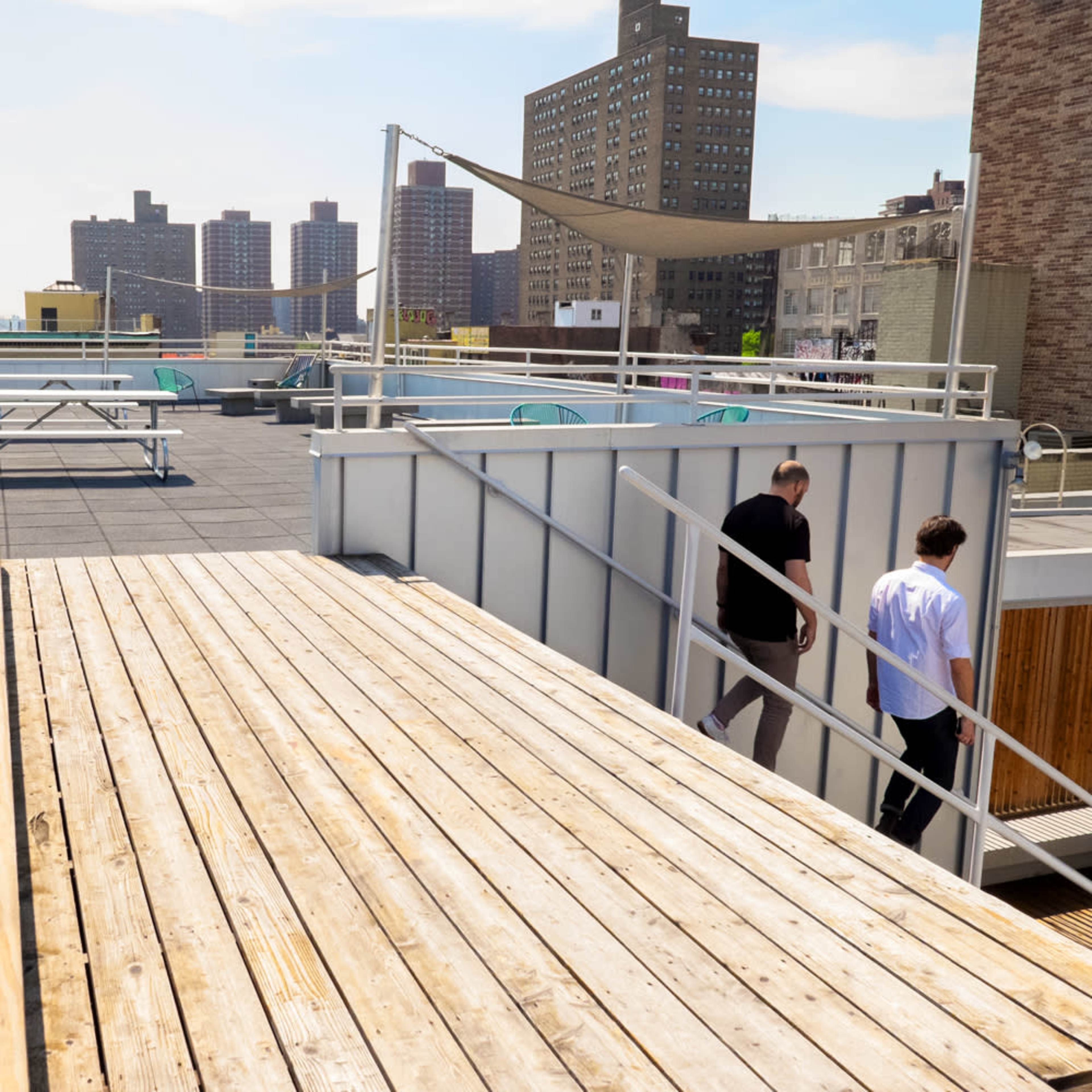 Two people walk along a wooden deck on a rooftop surrounded by buildings.