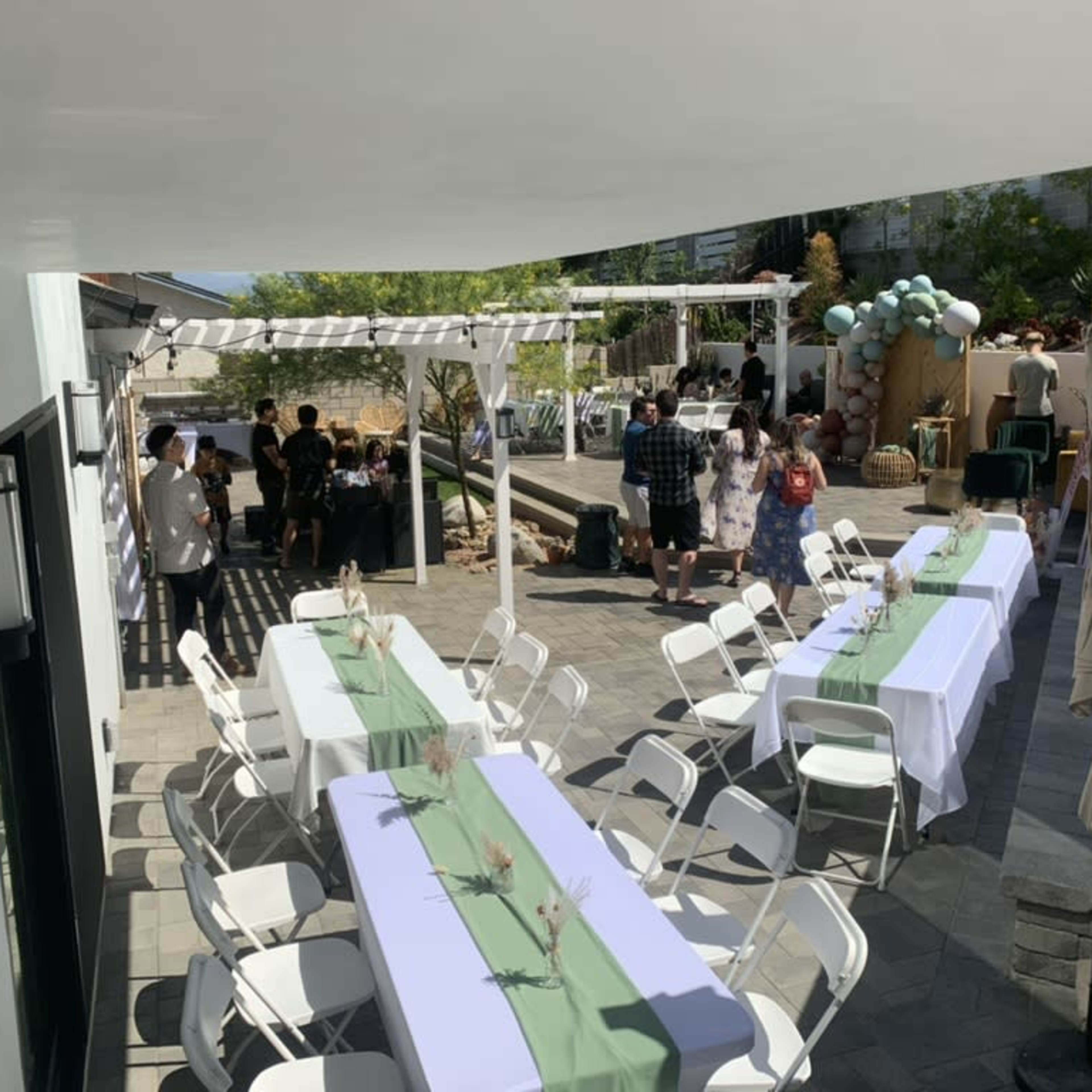 A patio area is set up for an event with rows of white tables and chairs, and guests mingling in the background.