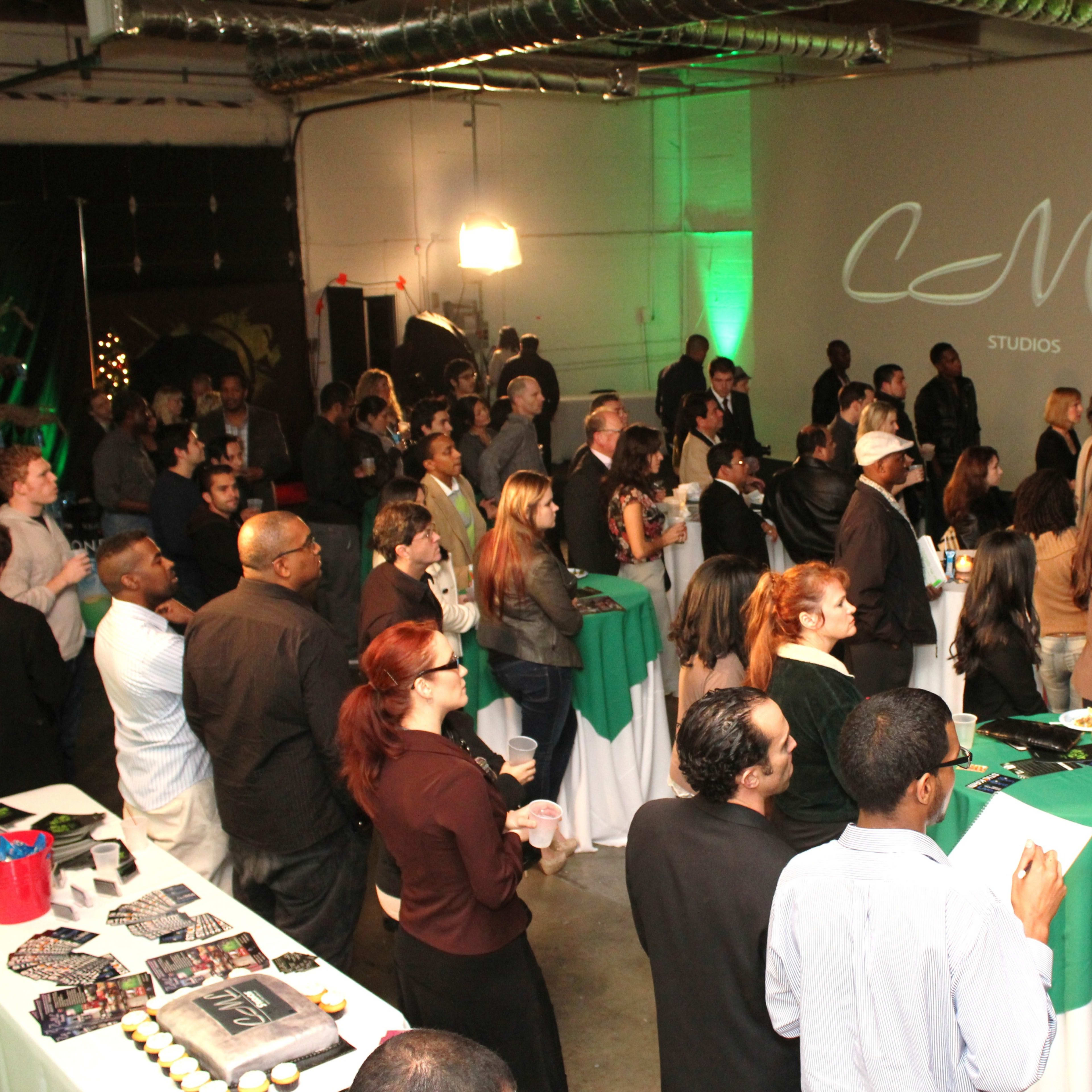 A large group of attendees stands in a studio space, facing a presentation screen in an event setting with tables covered in green and white.