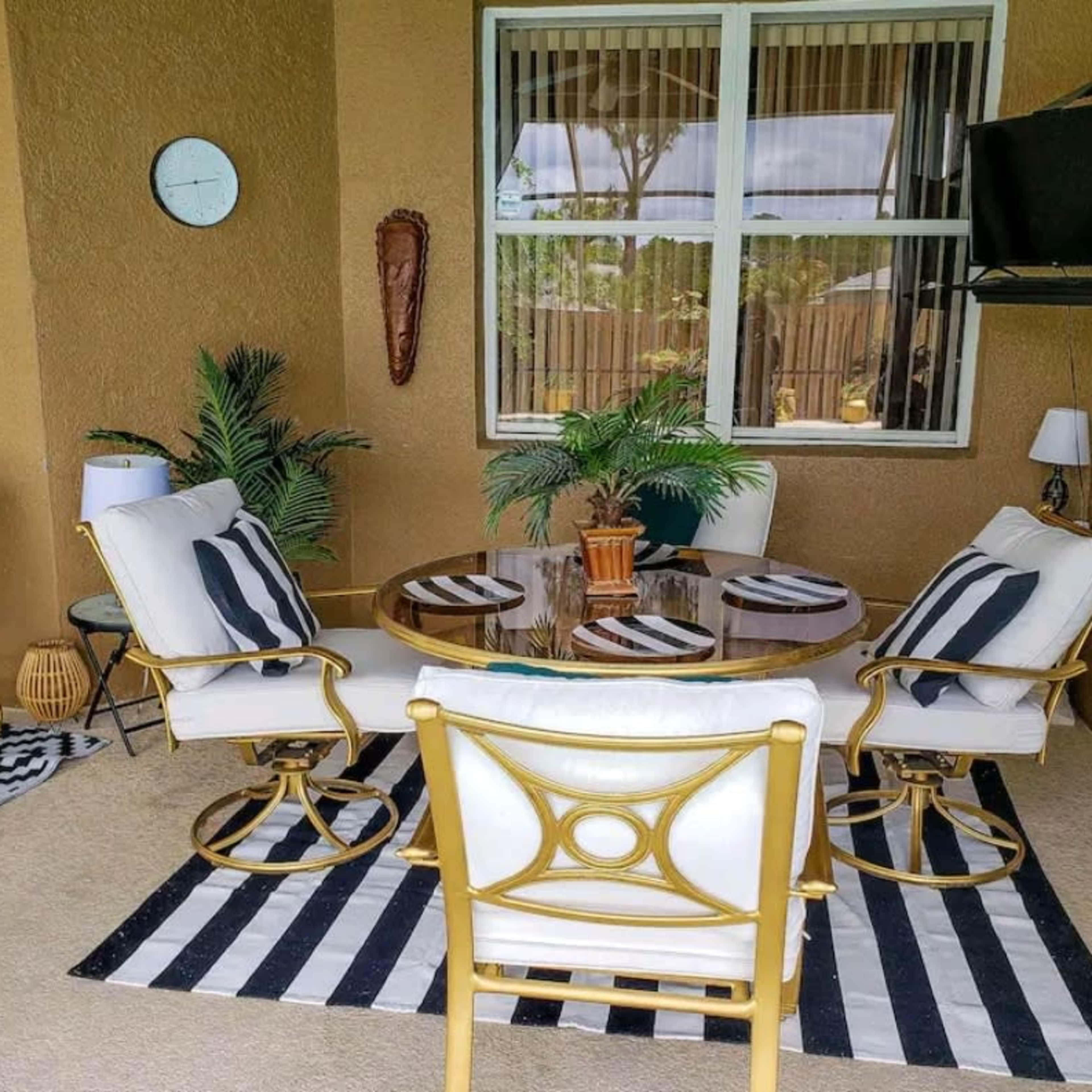 A cozy outdoor patio featuring a round glass table surrounded by white and black chairs, with a patterned rug underneath and several decorative plants.