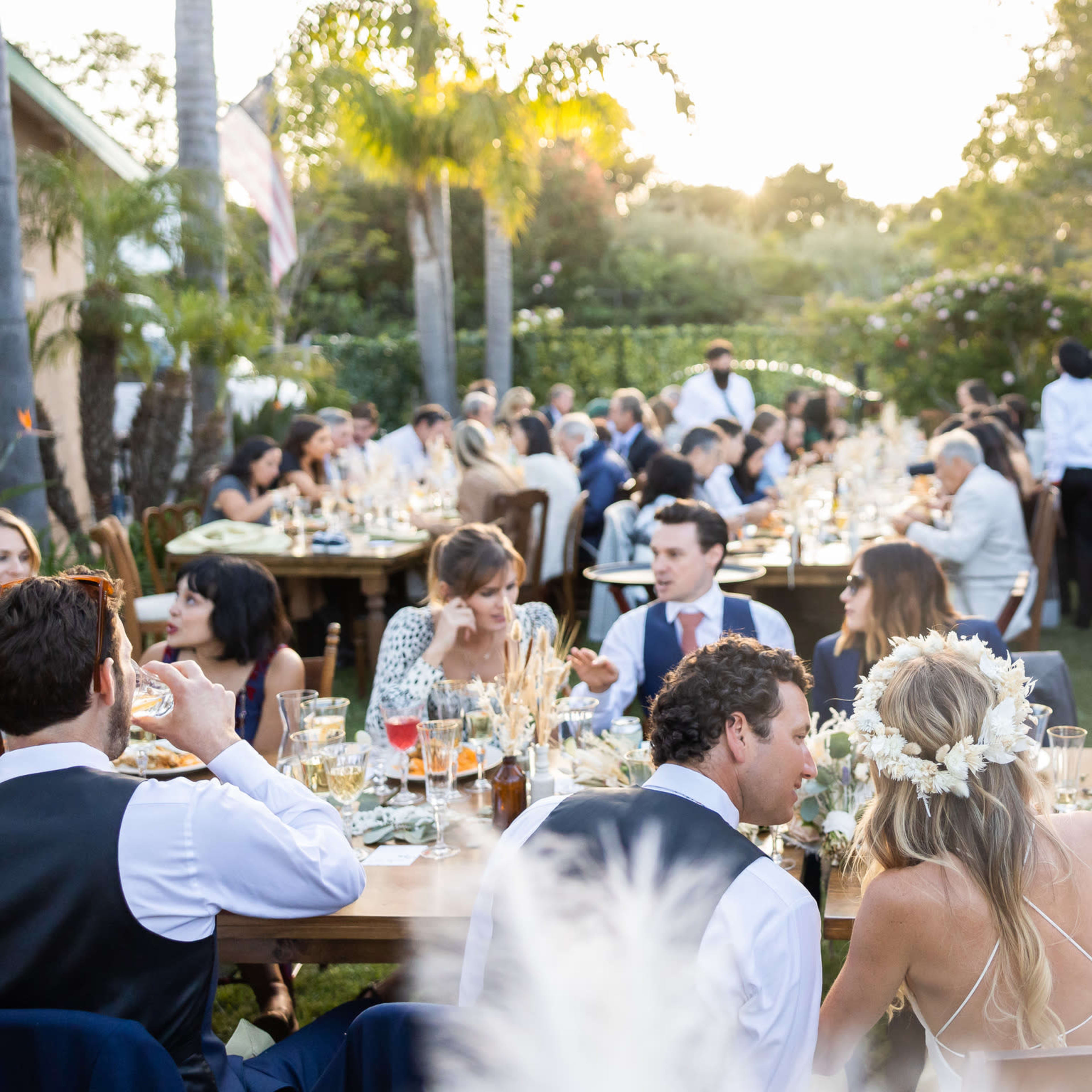 A large outdoor dinner party is set up with guests seated at long tables, enjoying their meals in a garden adorned with trees and soft lighting.