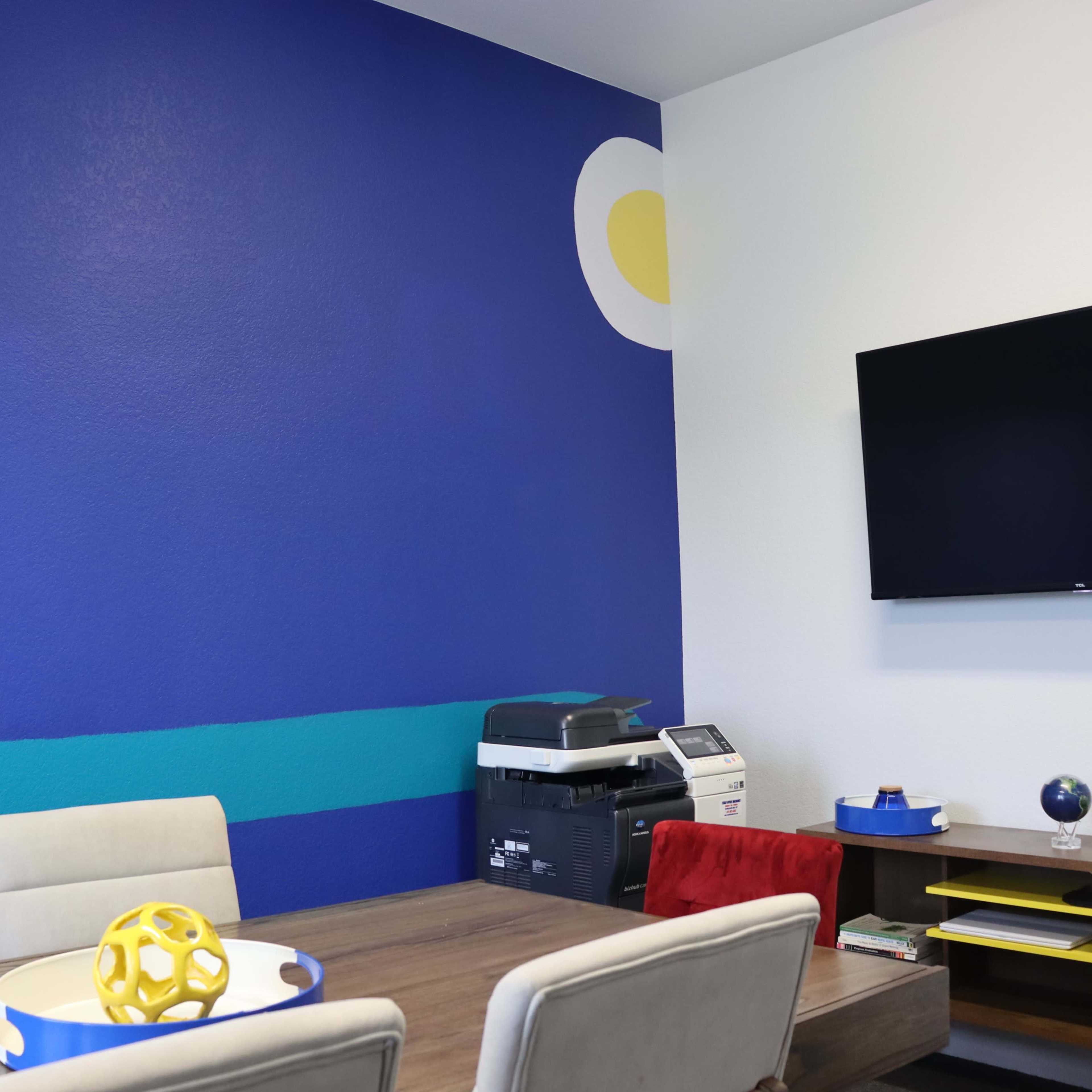 A conference room features a blue accent wall with a yellow circle, a wooden table with beige chairs, and a wall-mounted television.