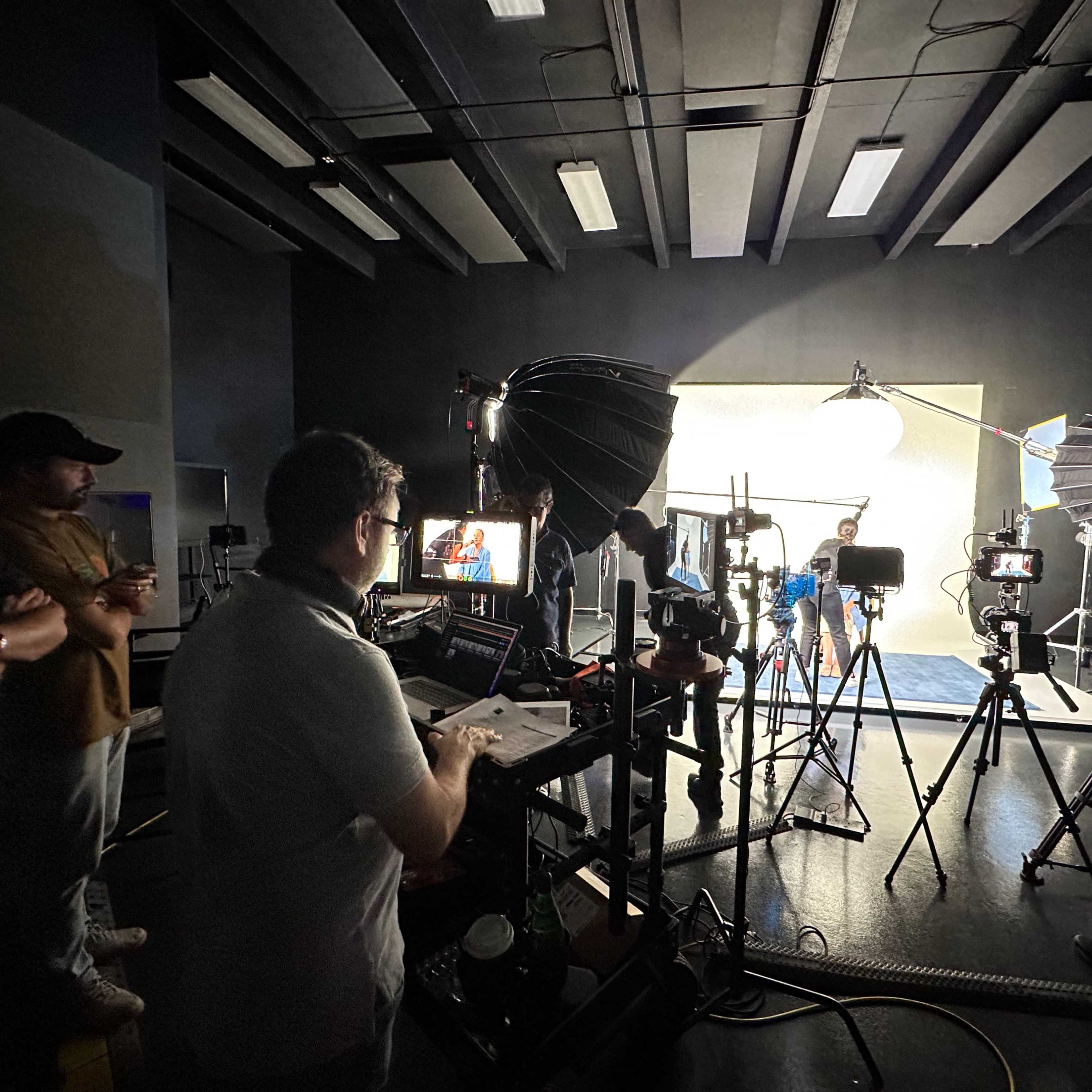 A film crew sets up lighting and equipment in a studio while a model poses against a backdrop.