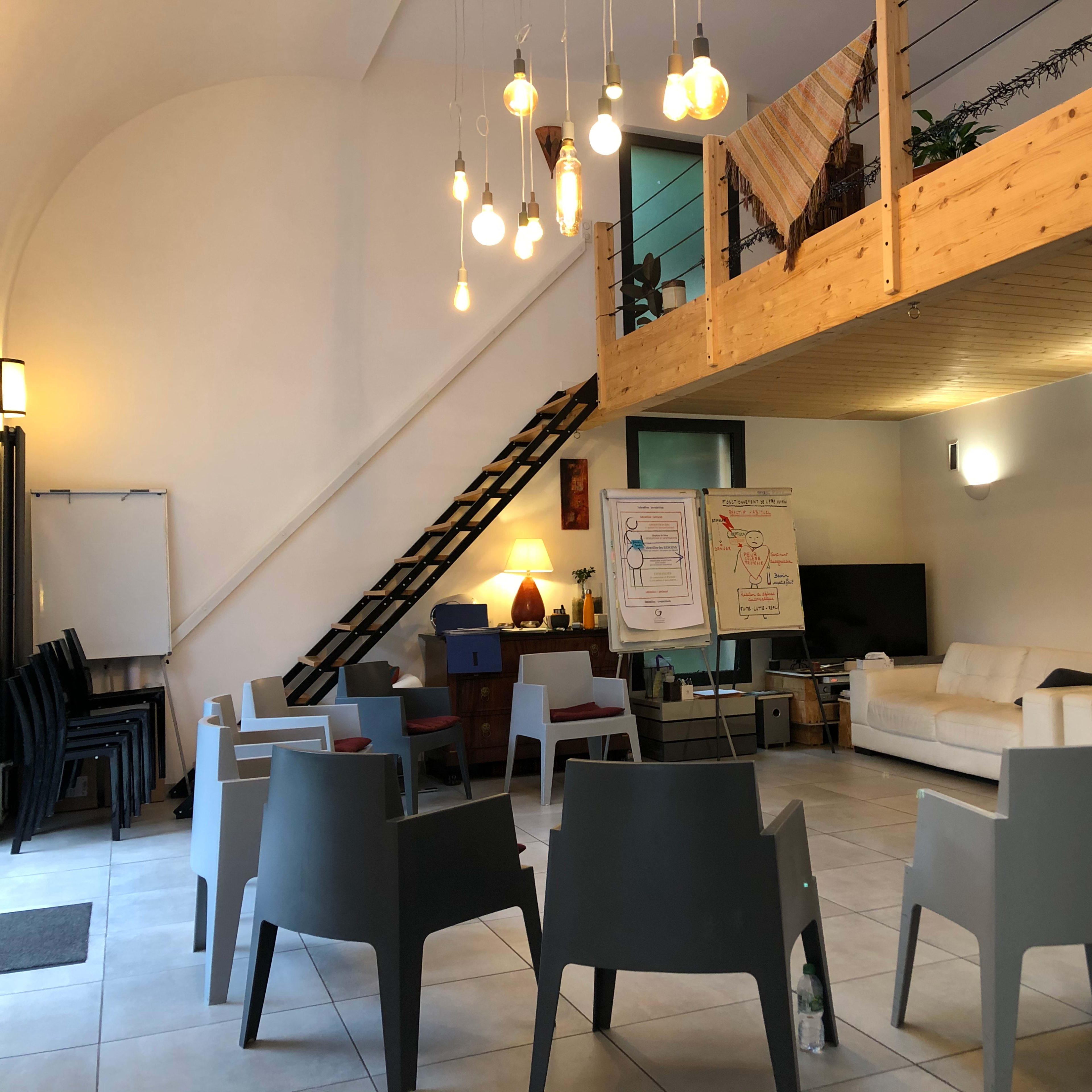 An interior room with a seating area arranged in a circle, a staircase leading to a wooden loft, and a presentation board beside a television.