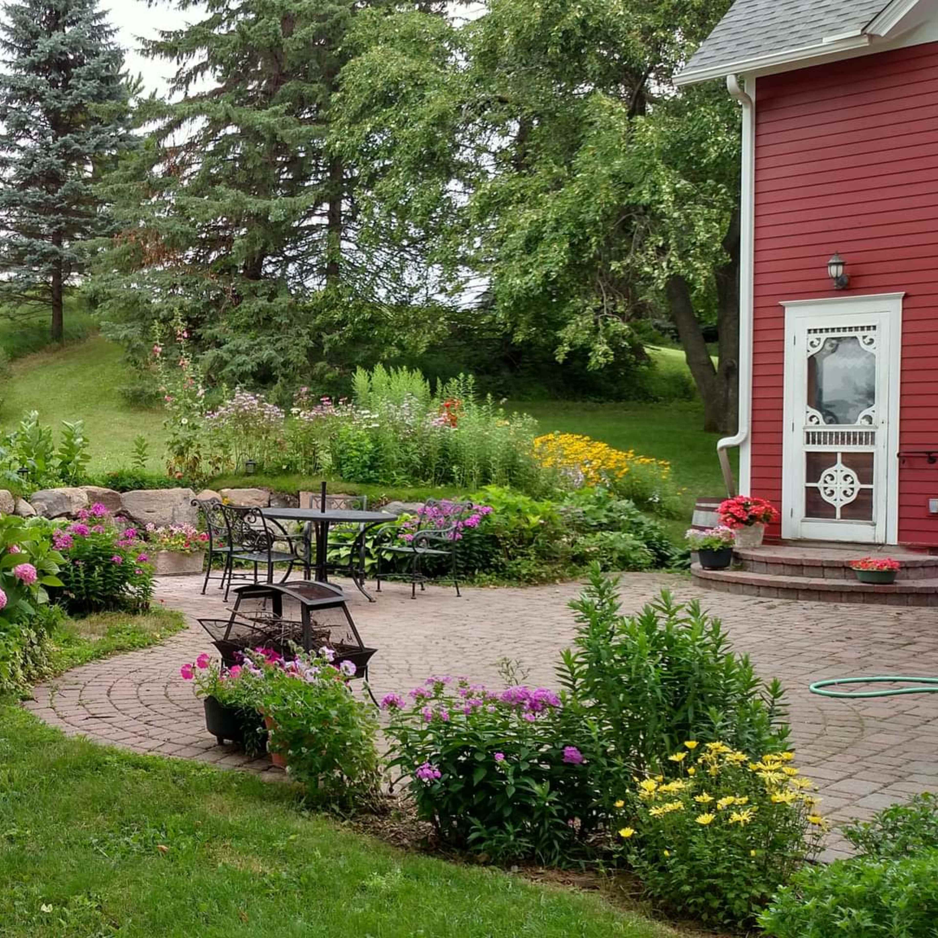 A landscaped garden featuring a stone pathway, a seating area, and a variety of colorful flowers surrounding a red house and a white building in the background.