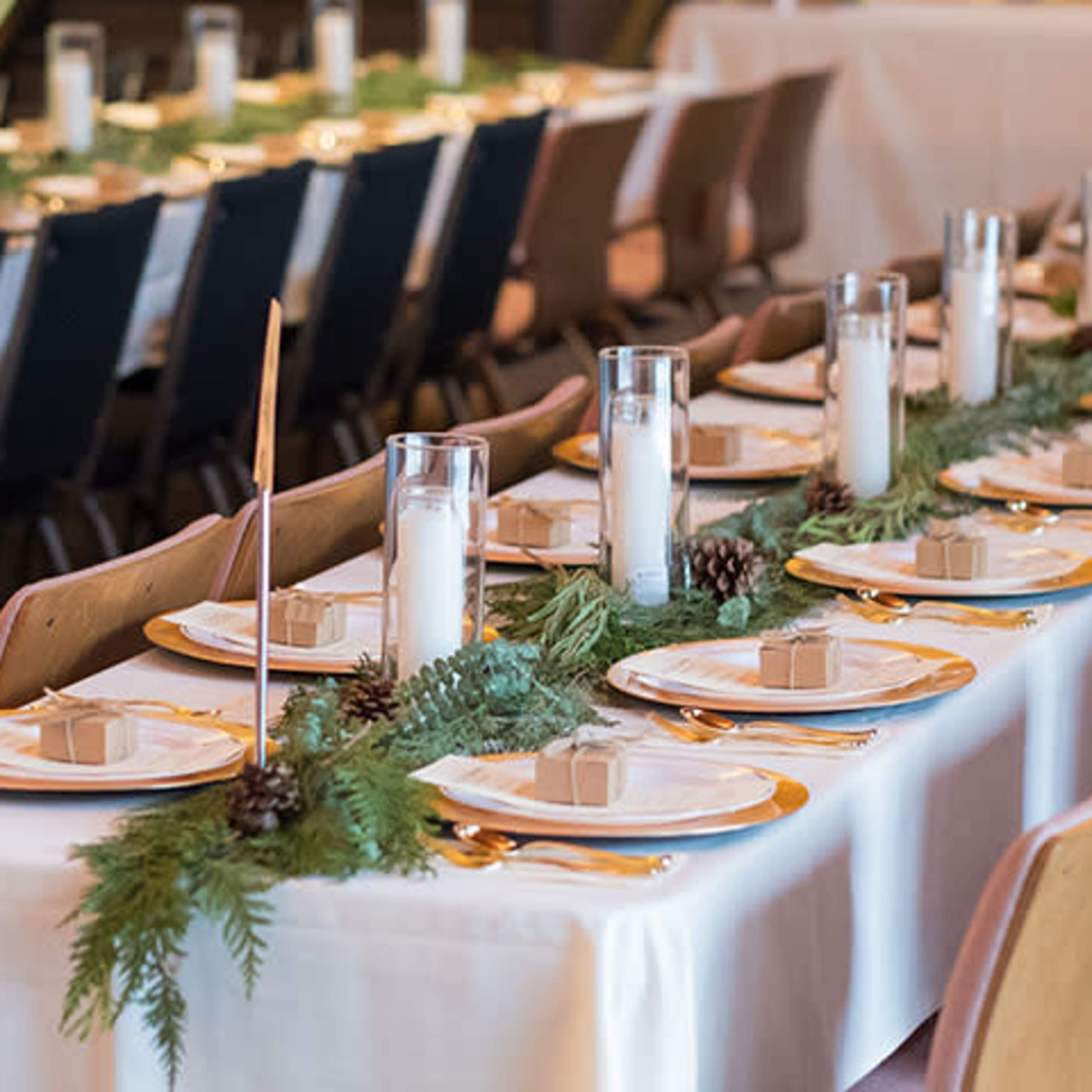 A long banquet table is set for a formal dinner with white tablecloths, plates, candles in glass holders, and greenery arranged down the center.