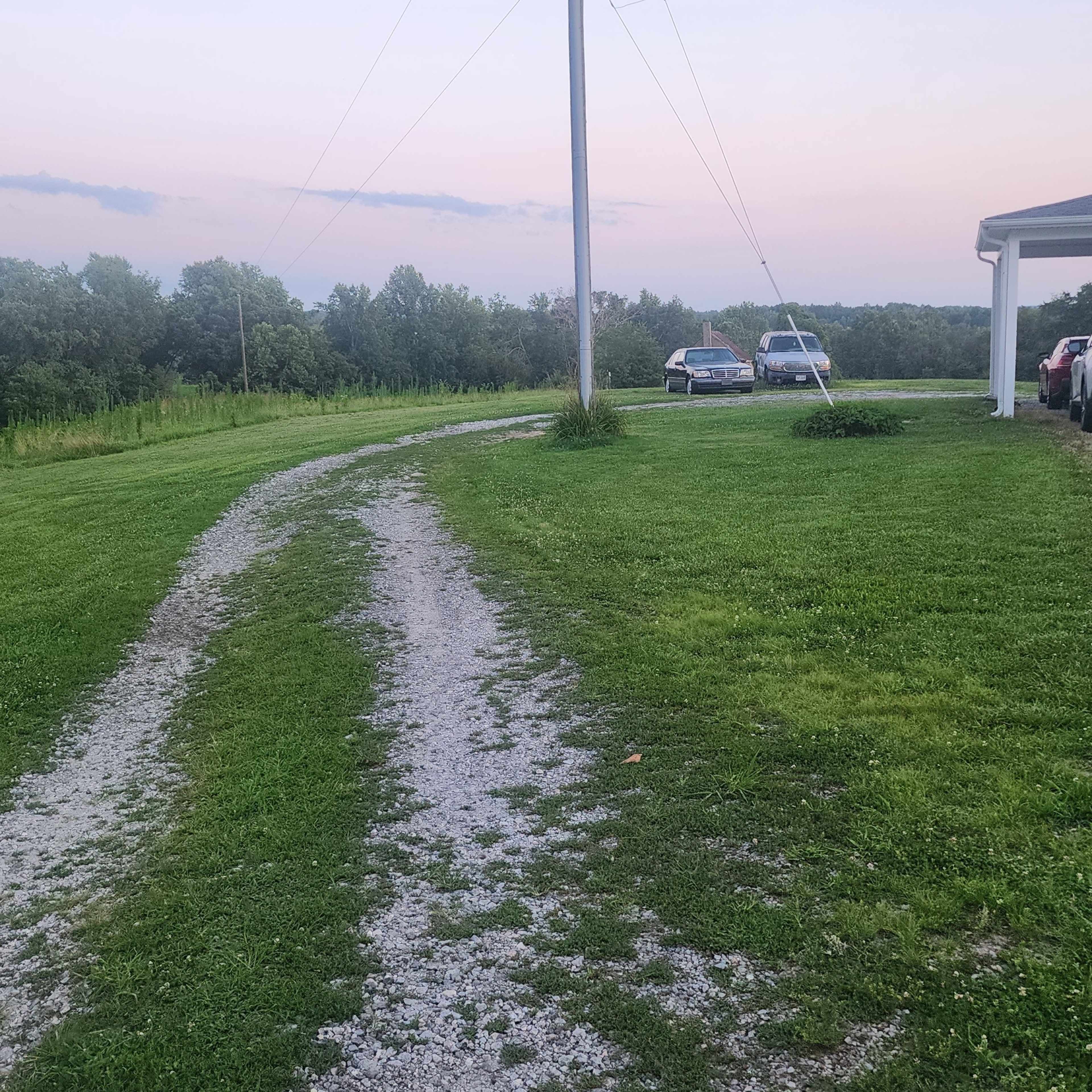 A gravel driveway winds through green grass towards two parked cars near a utility pole, with trees and a sunset sky in the background.