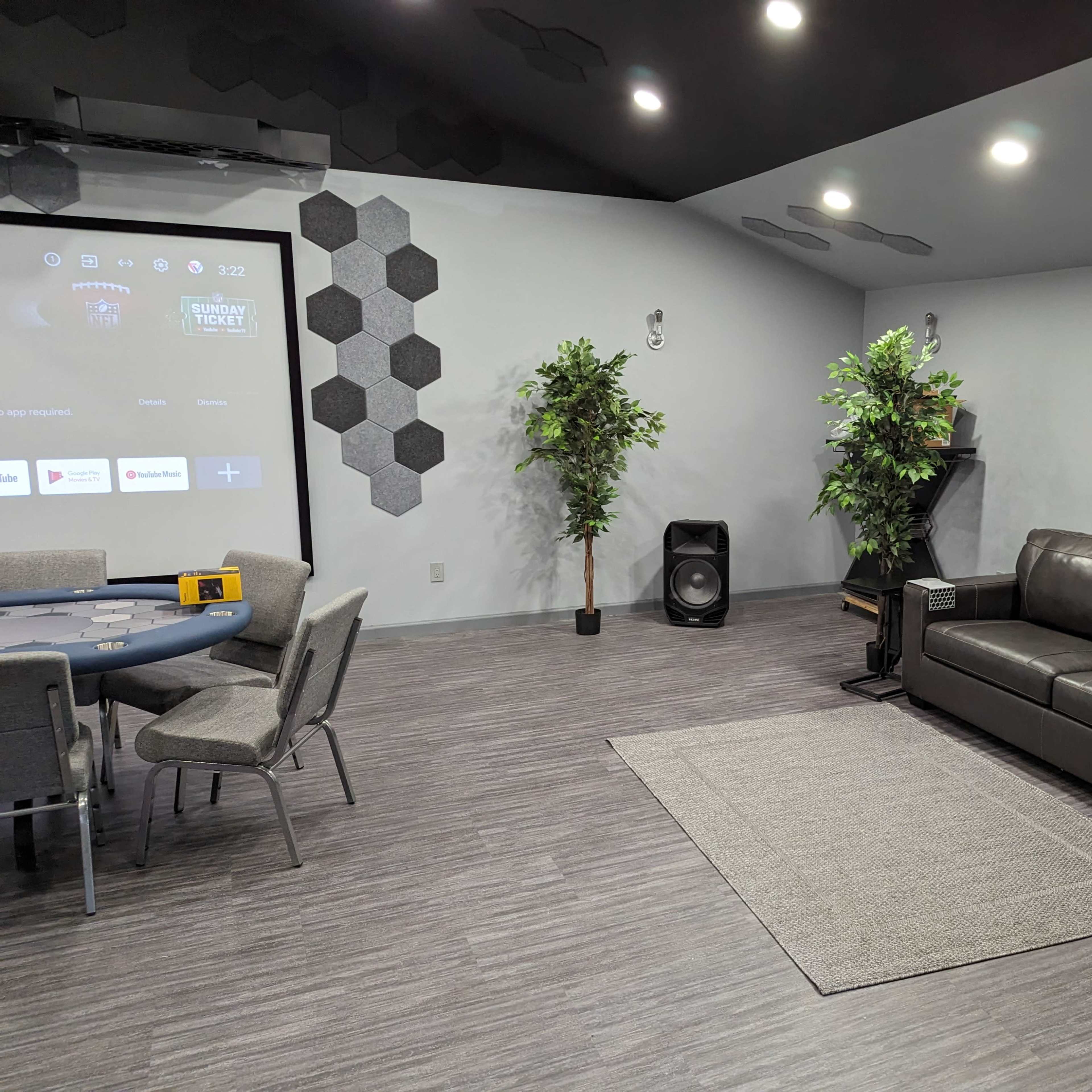 A modern room featuring a poker table, a grey sectional sofa, two artificial plants, and a projector screen on the wall.