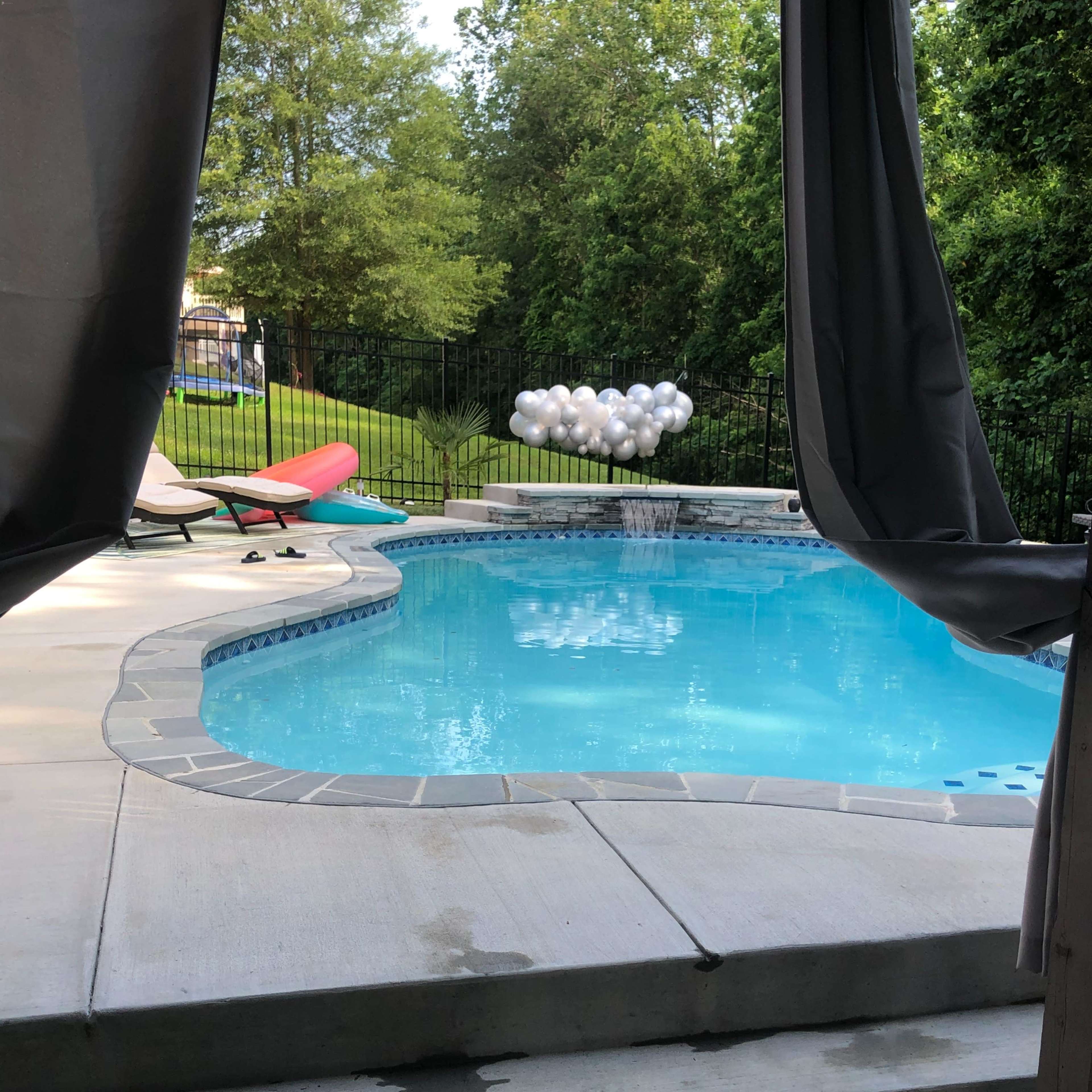 A swimming pool surrounded by a concrete patio, with a set of lounge chairs and a cluster of white balloons visible in the background.