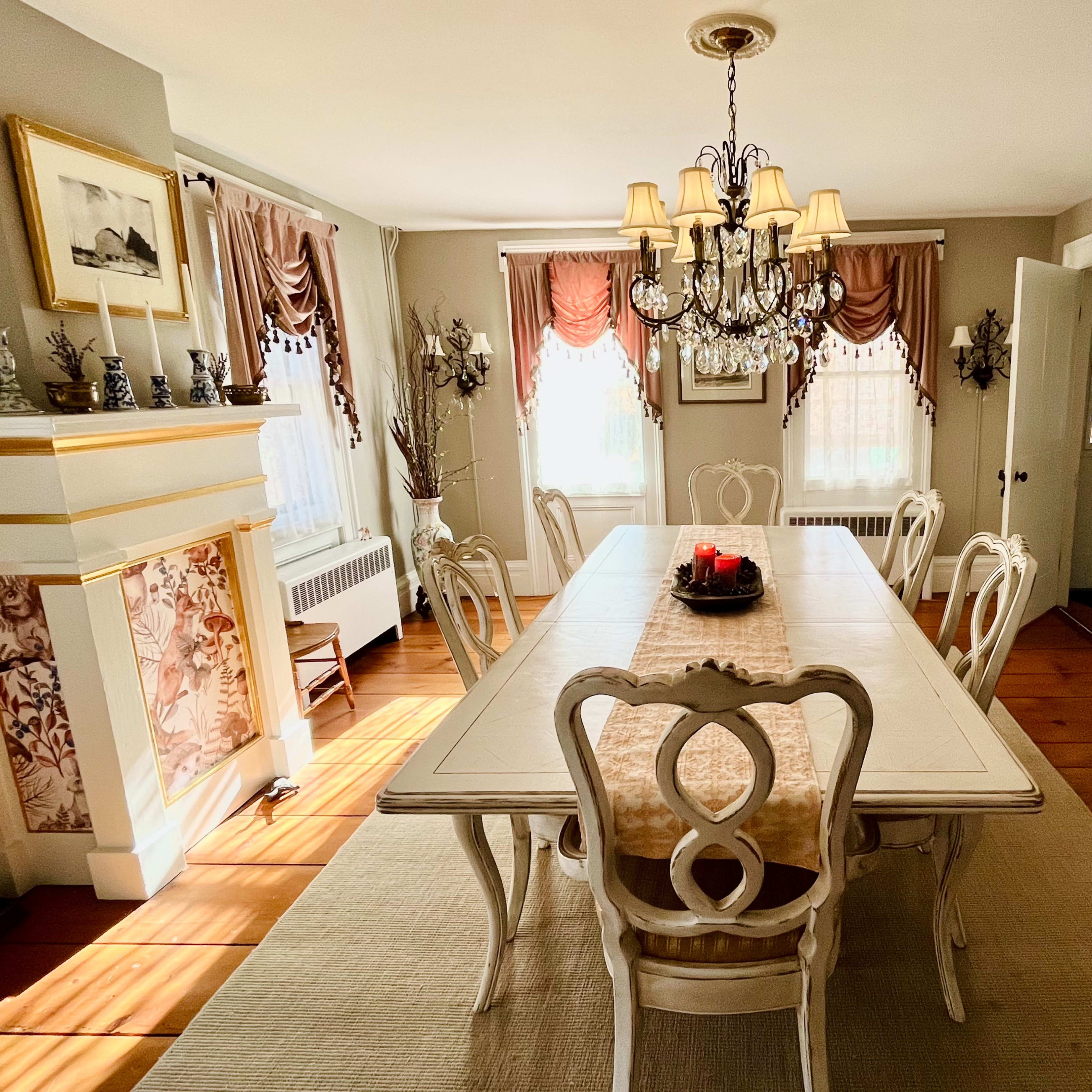 A well-lit dining room features a long table surrounded by ornate chairs, a chandelier overhead, and decor including curtains and wall art.