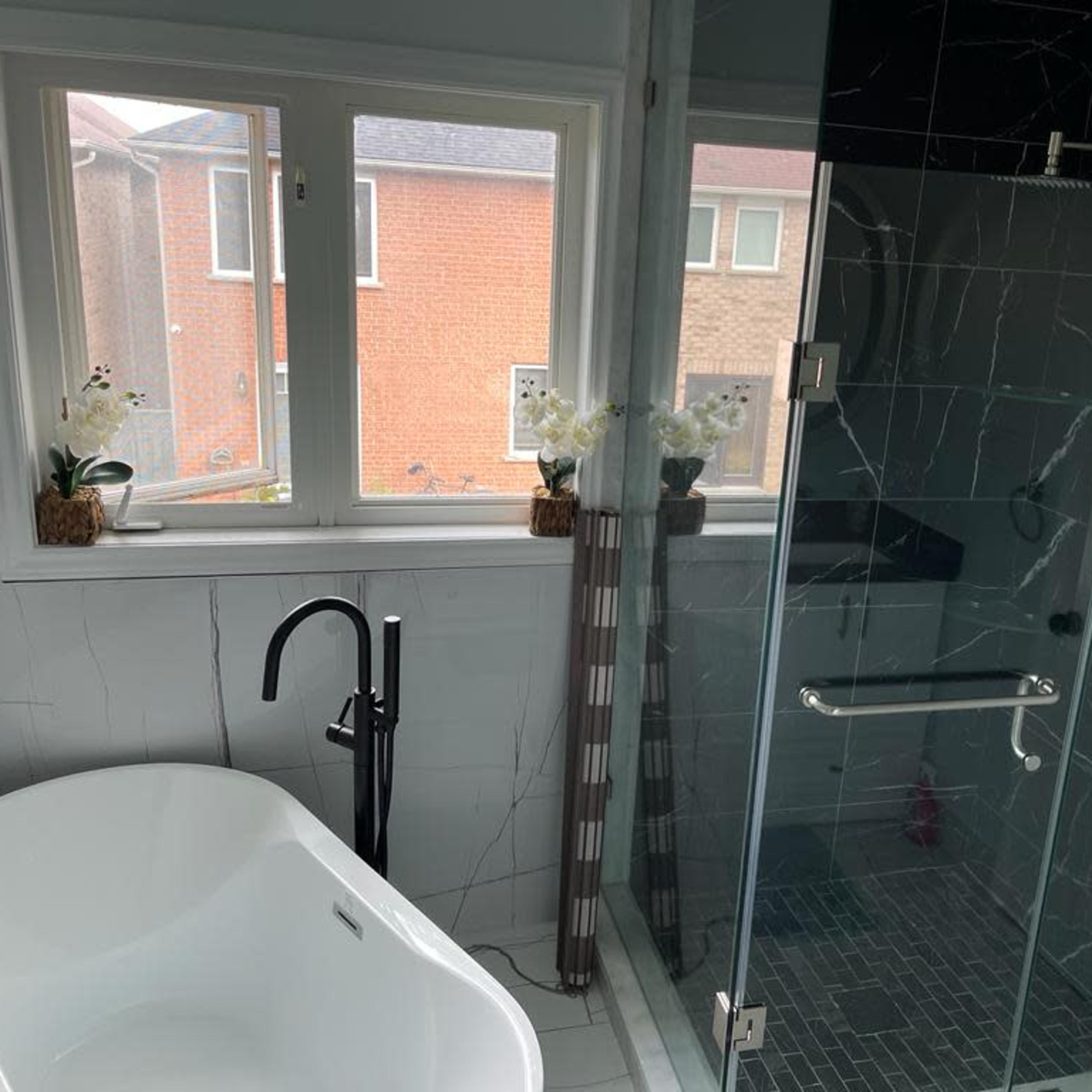 A bathroom featuring a freestanding white bathtub, a glass-enclosed shower, and large windows with potted flowers on the sill.