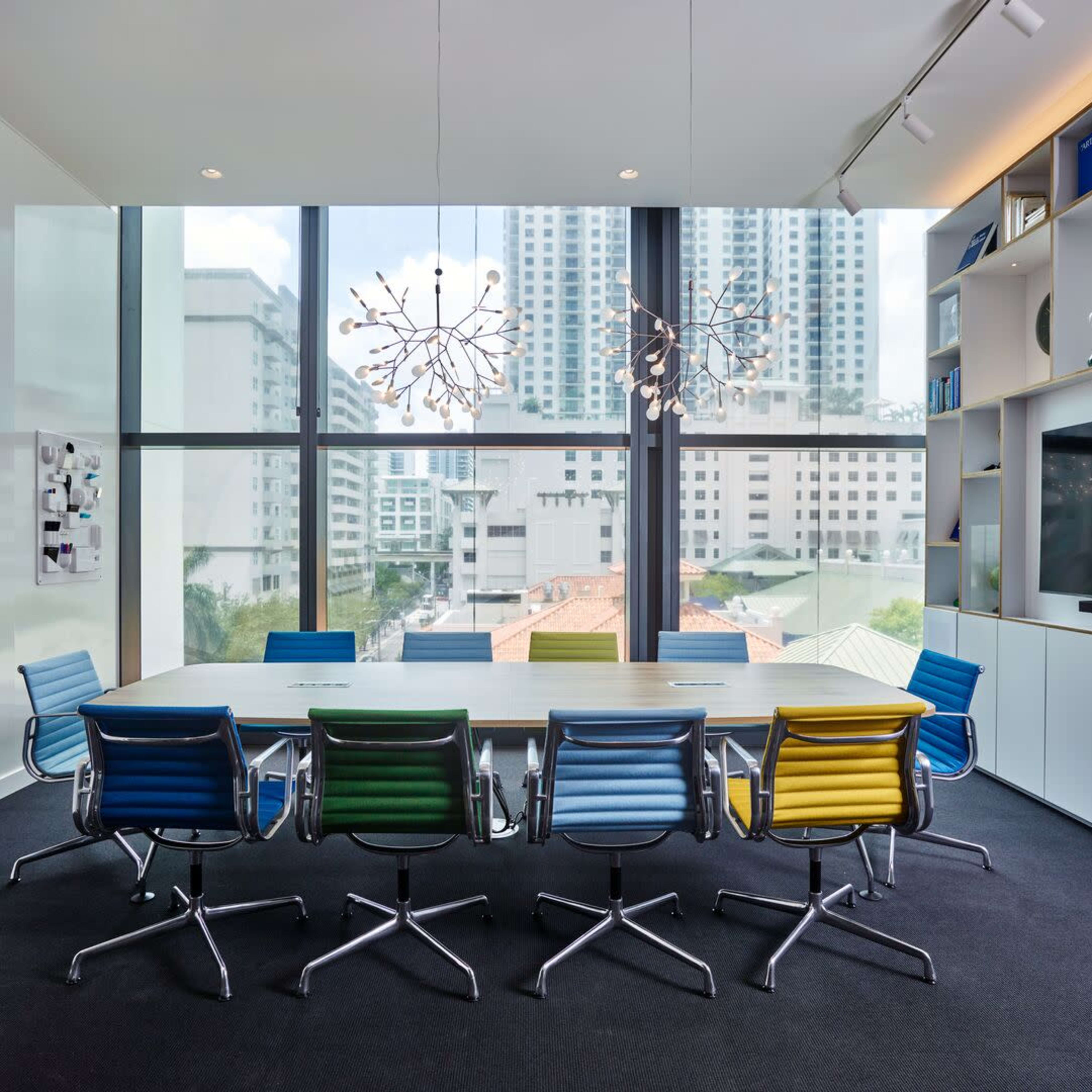 A modern conference room features a long table surrounded by colorful chairs and large windows showcasing a city view.