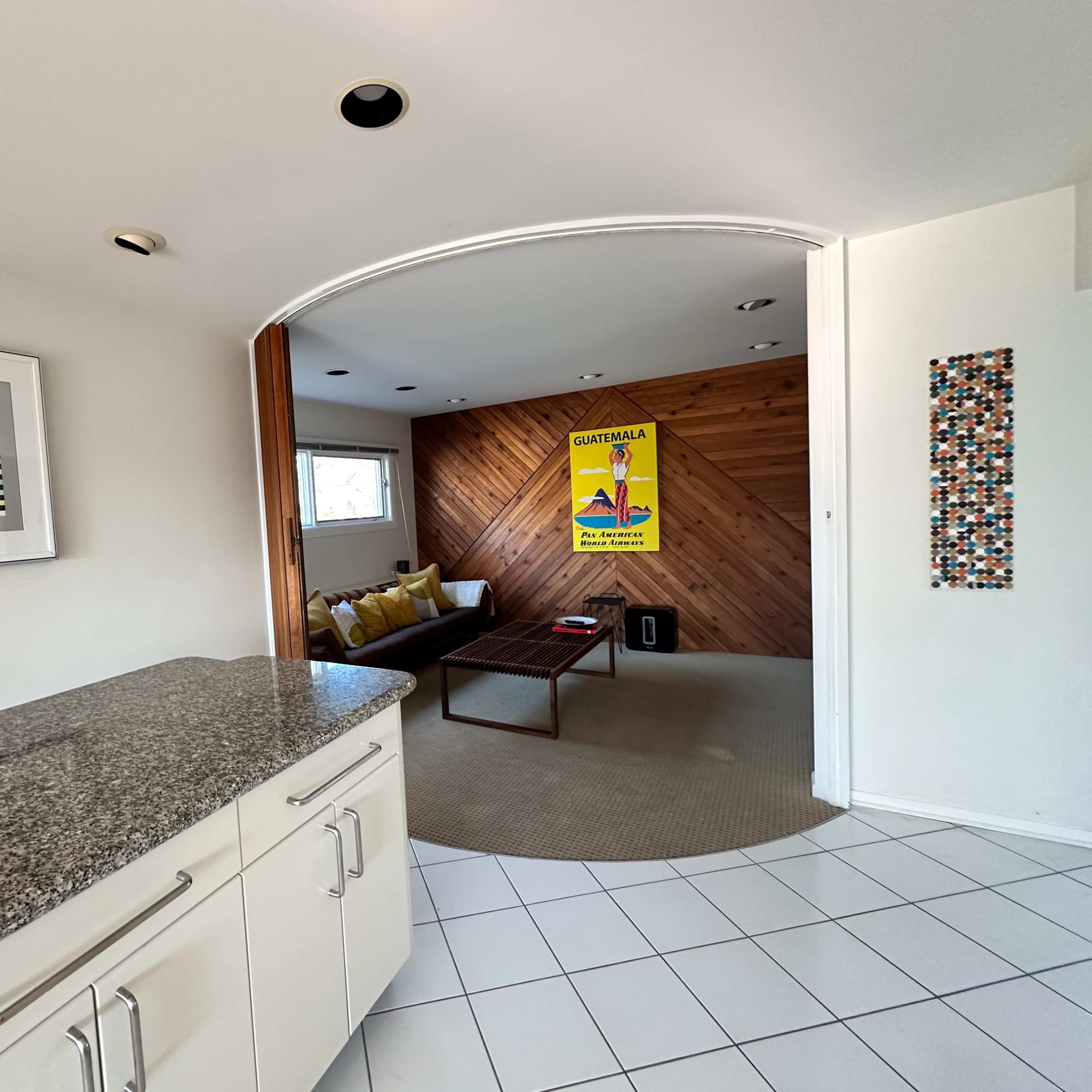 A modern kitchen with a granite countertop leading into a living area featuring a wooden accent wall and a colorful poster.