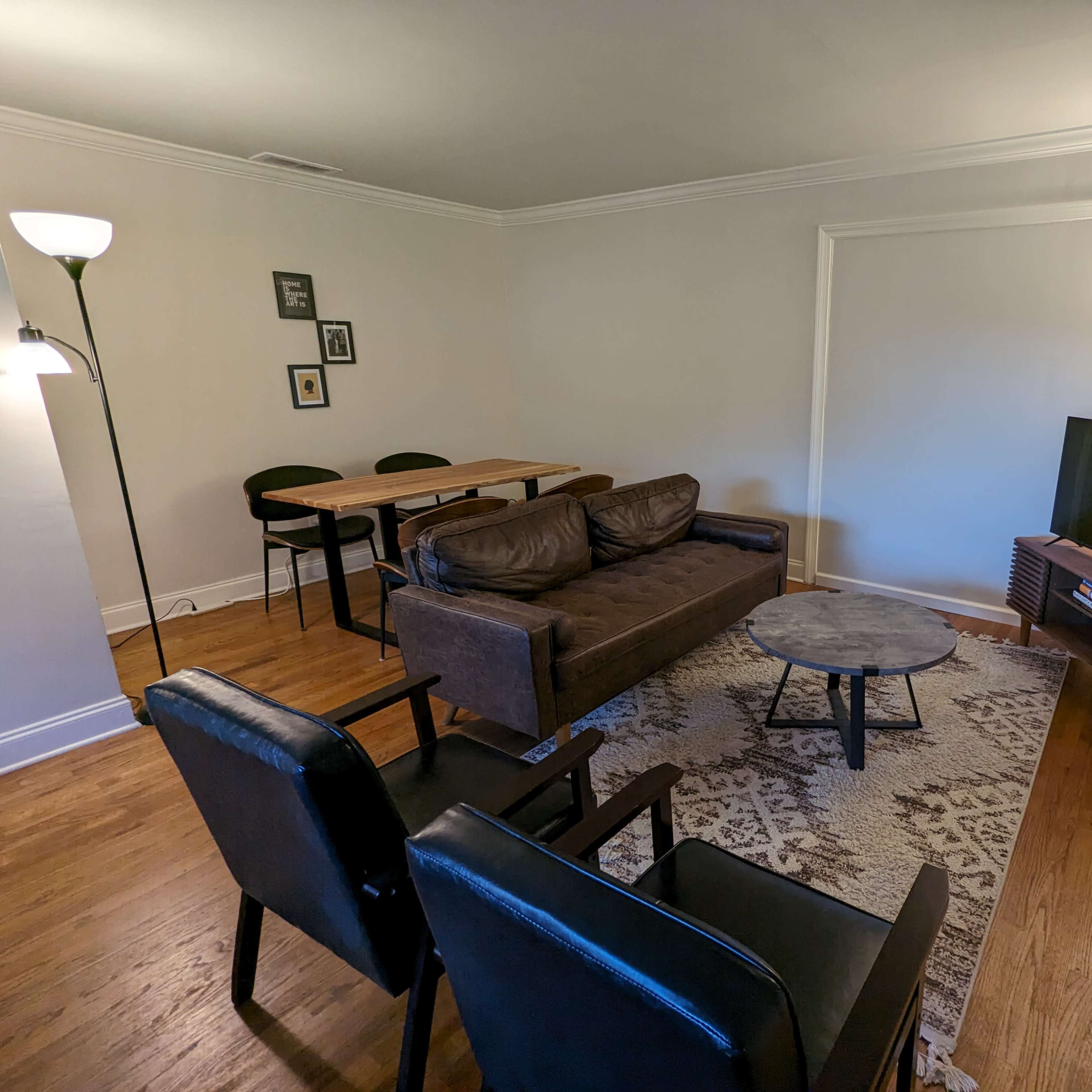 A living room features a brown sofa, two black chairs, a round table, a TV on a stand, and a patterned area rug on hardwood flooring.