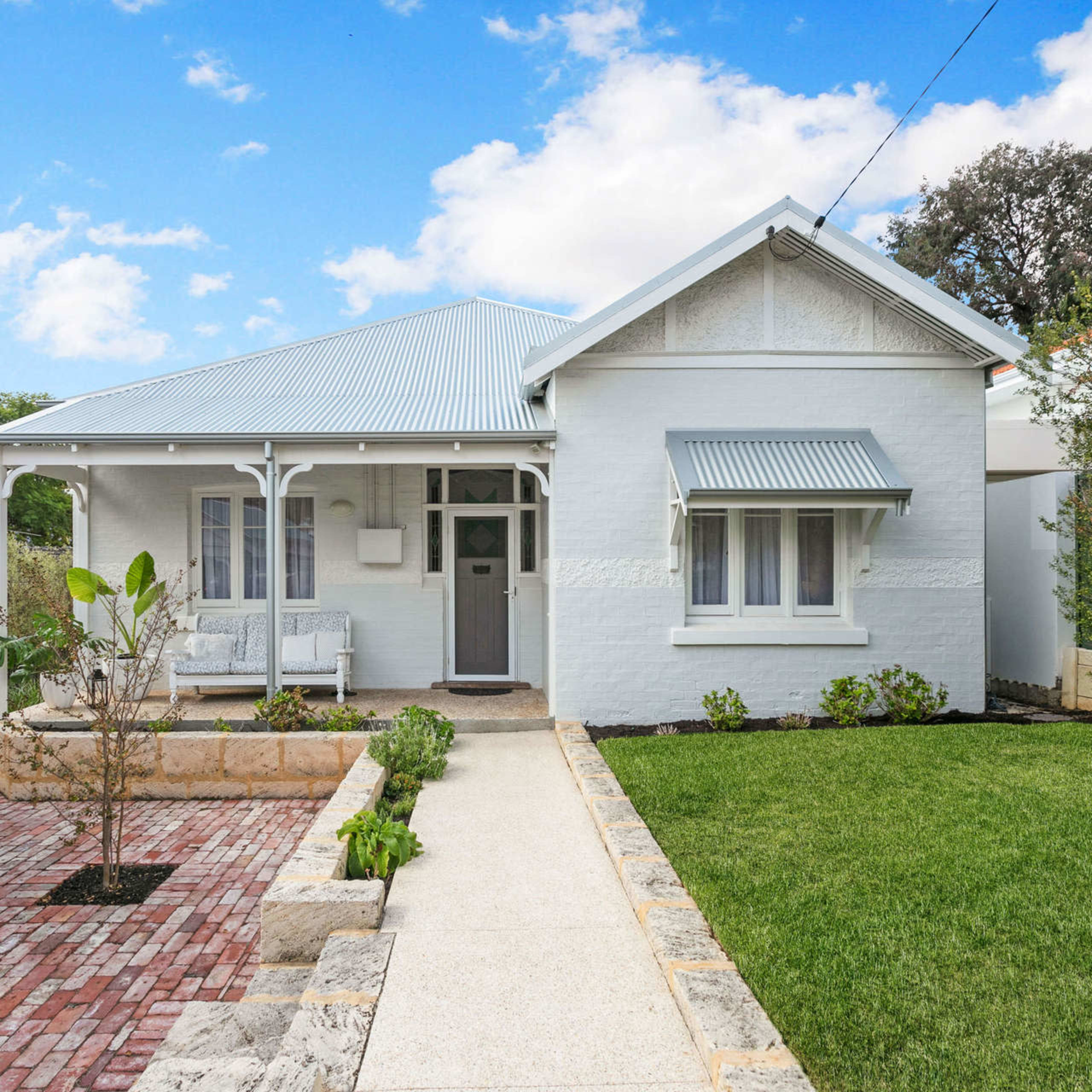 A white cottage with a light blue tin roof, featuring a front porch and a landscaped yard with brick pathways and greenery.