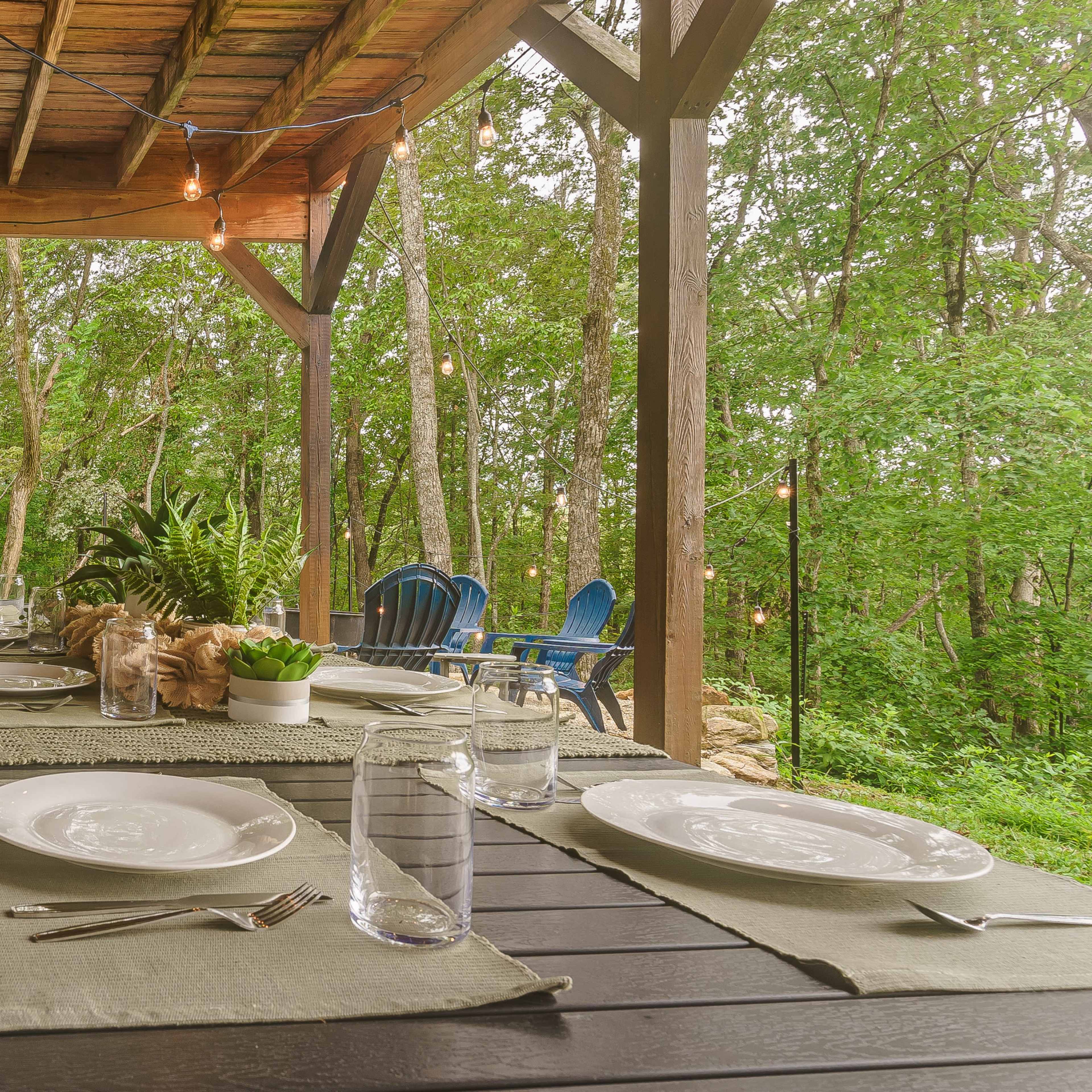 A dining table is set under a wooden porch surrounded by trees, with plates, glasses, and plants arranged neatly on the table.