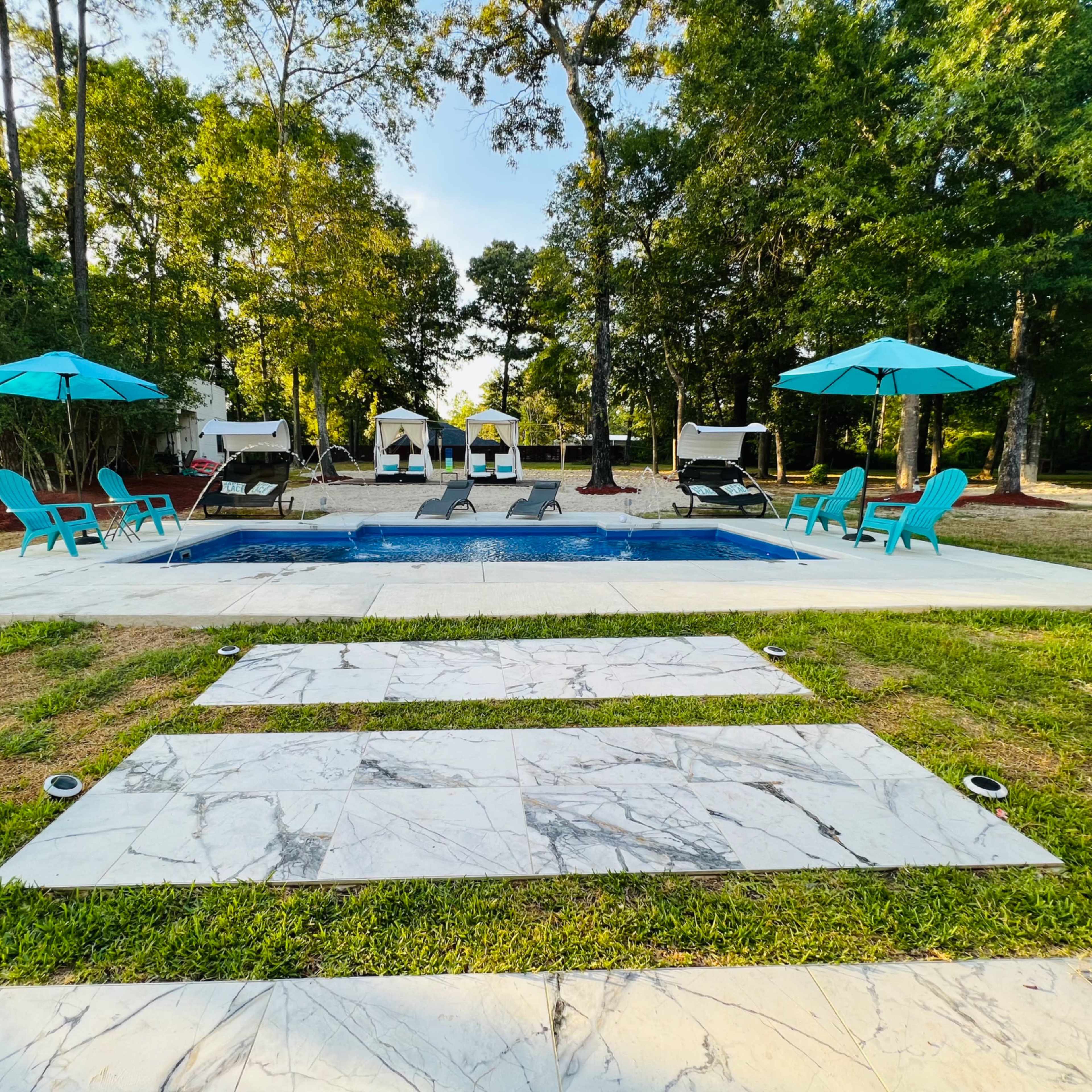 A backyard pool area with lounge chairs and umbrellas, surrounded by trees and a grassy lawn.
