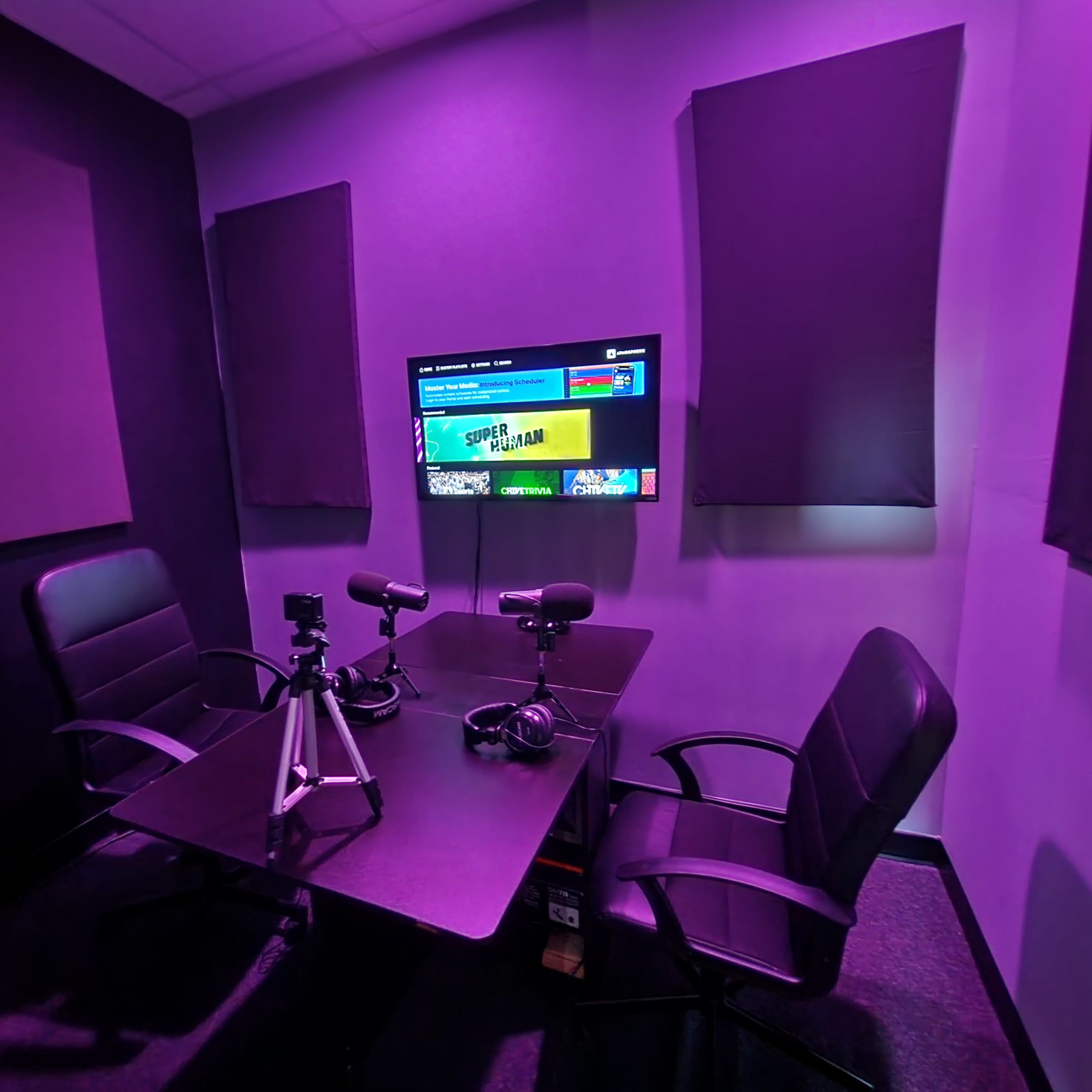 A small recording booth with two black chairs, a table, microphones on tripods, and a television displaying a menu, all set against purple walls with soundproof panels.