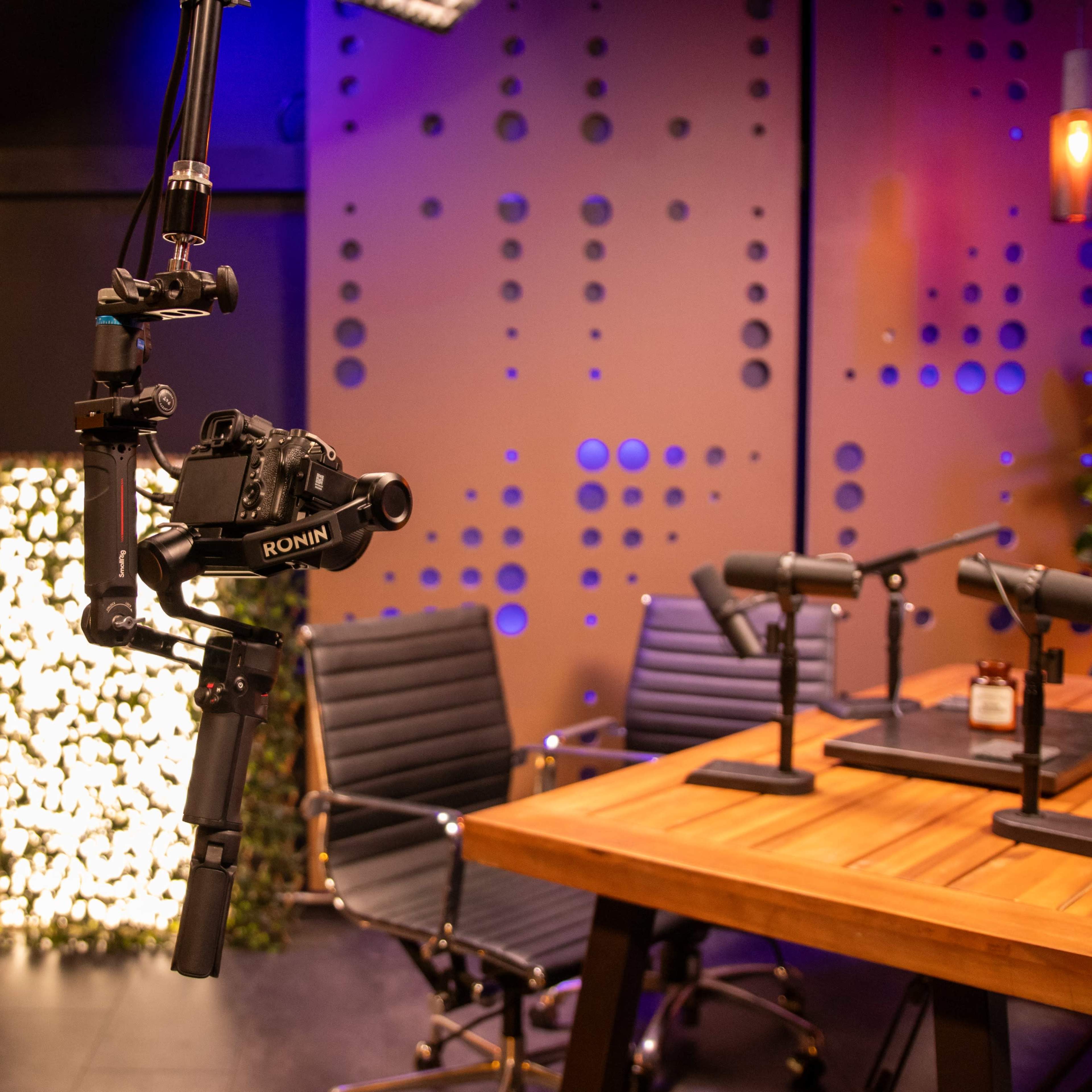A modern studio setup featuring a camera mounted on a boom arm, multiple microphones on a table, and a backdrop with patterned walls and greenery.