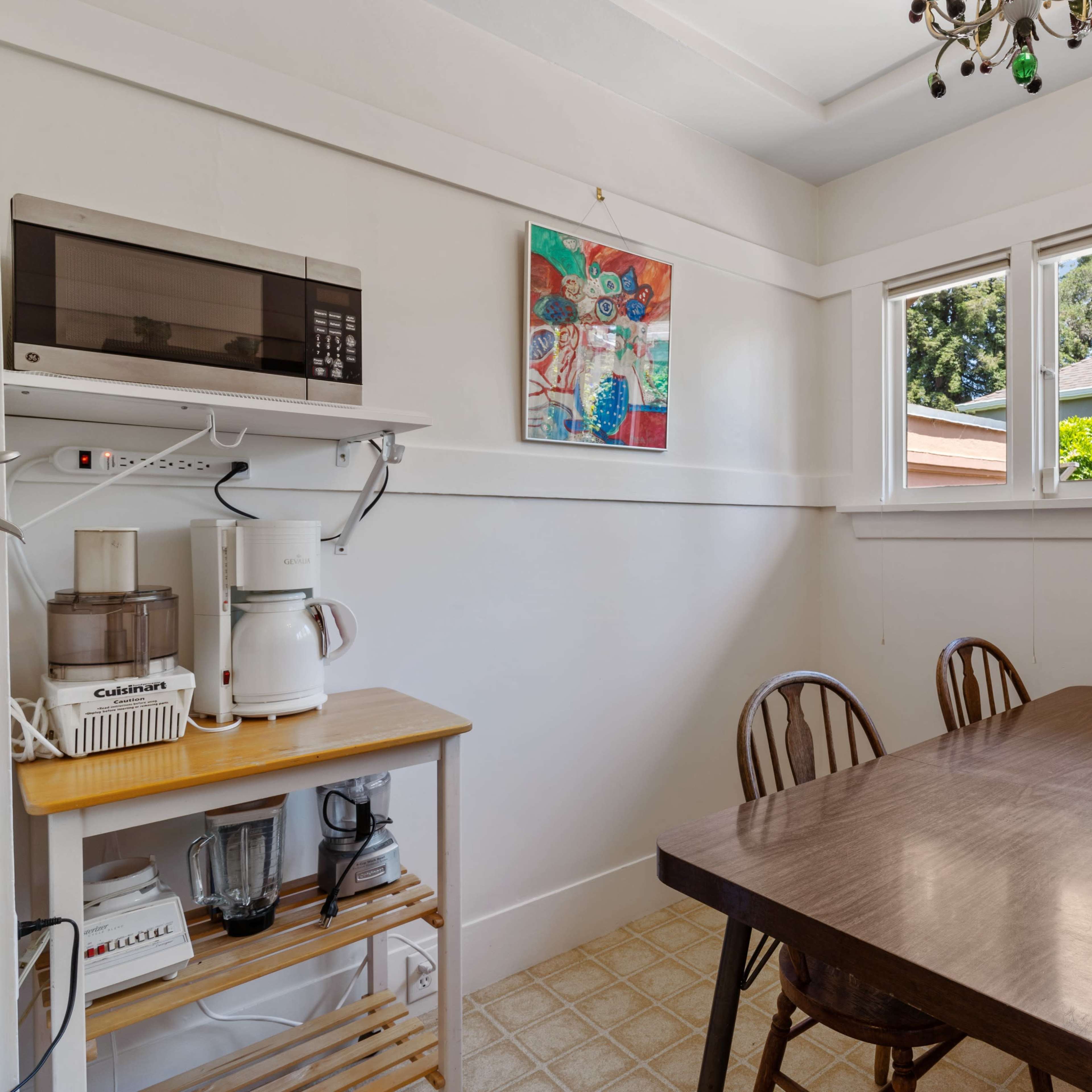 A small kitchen features a wooden dining table and chairs, with a microwave and appliances on a shelf, and windows providing natural light.