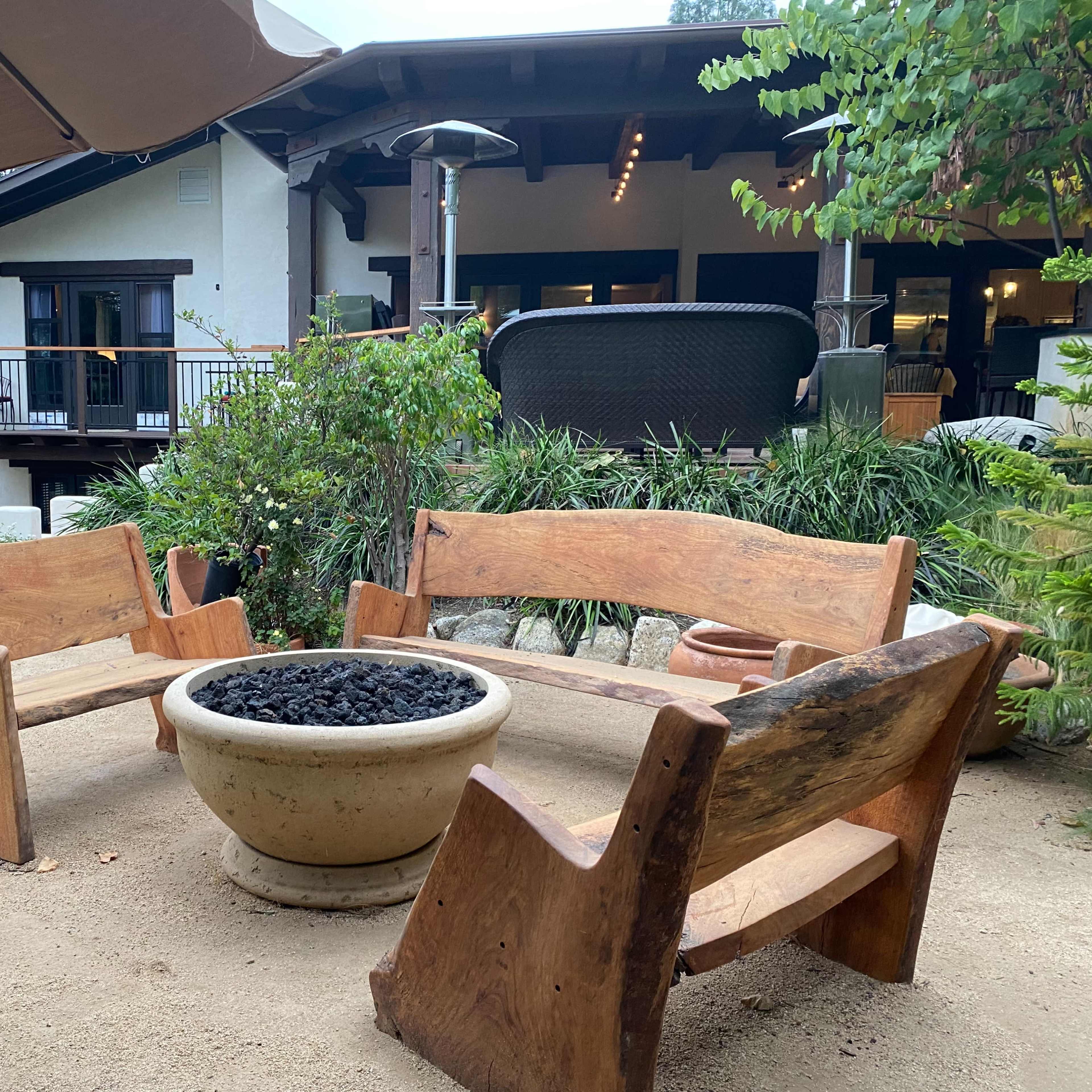 A circular seating arrangement made of wooden benches around a stone fire pit in a garden setting.