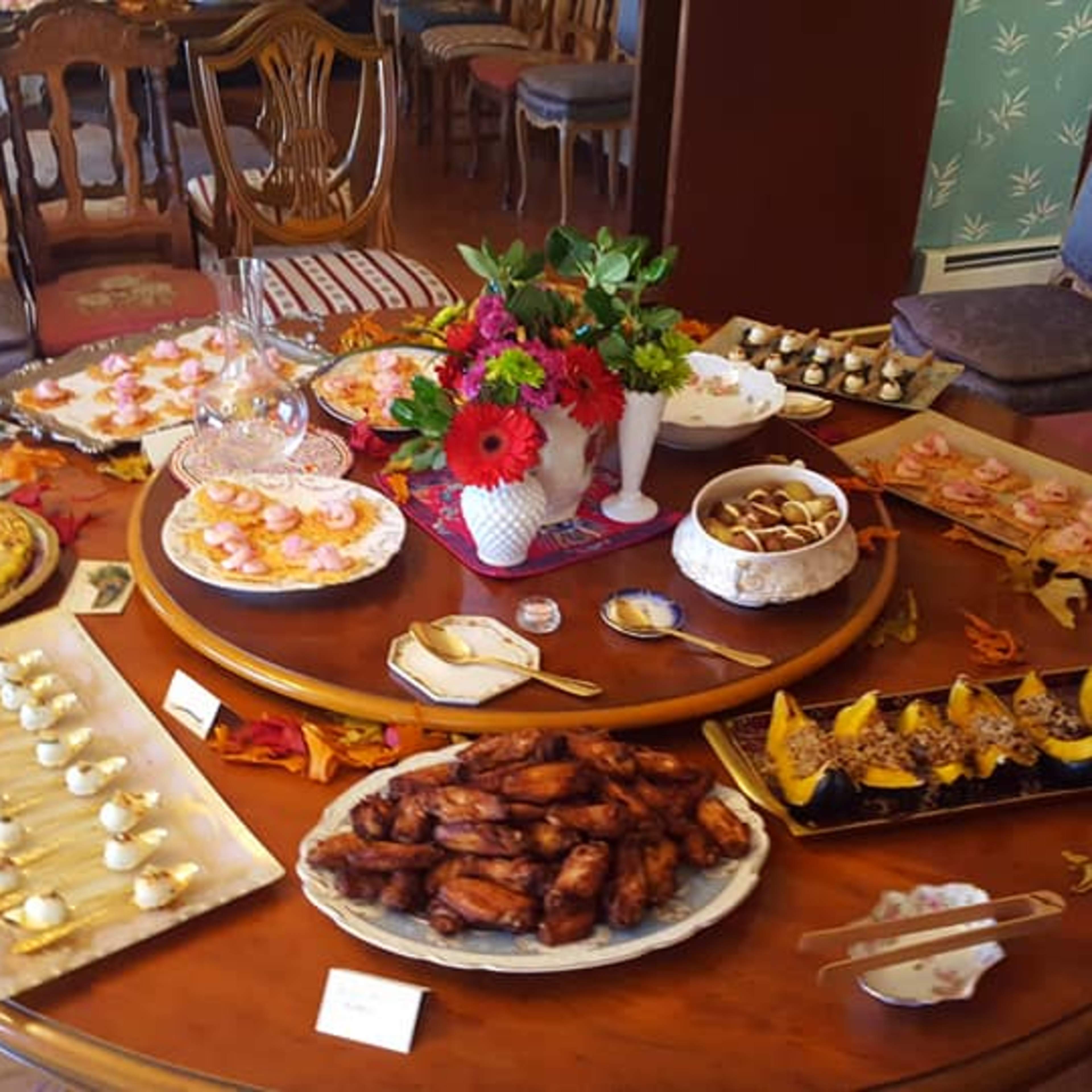 A round wooden table is set with an array of various dishes and desserts, including savory pastries, chicken wings, and beautifully arranged sweets, surrounded by decorated chairs.