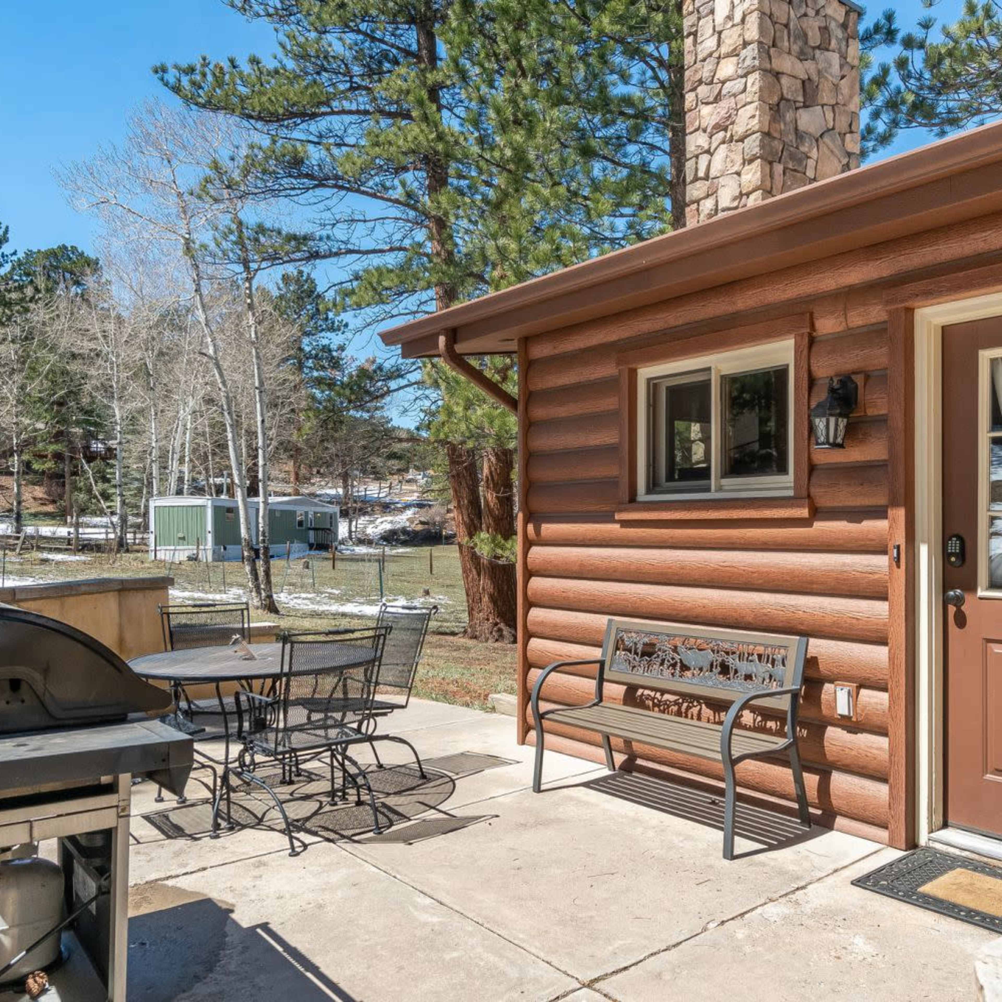 A log cabin features a patio with a grill and a table set for outdoor dining, surrounded by pine trees and patches of snow.