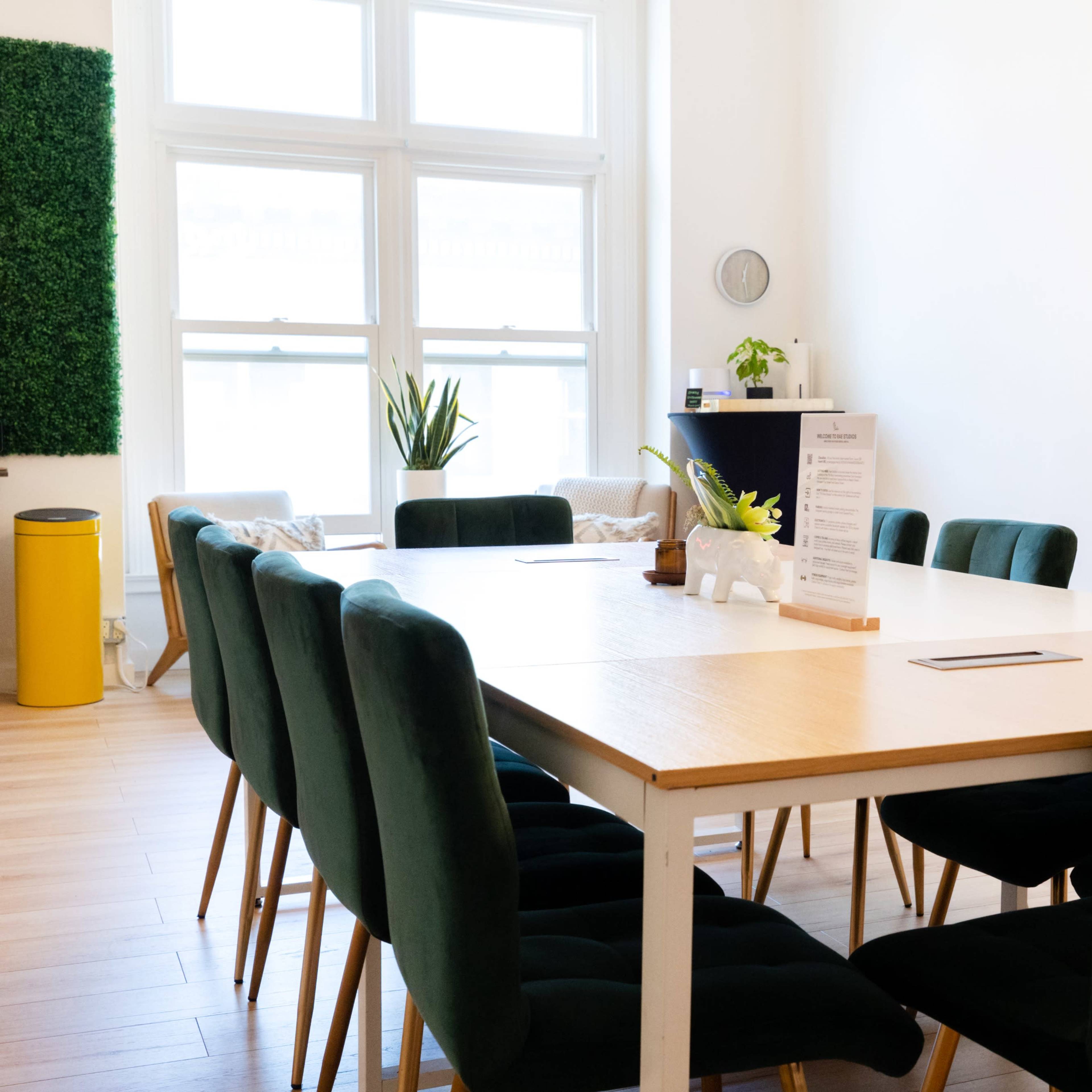 A long wooden table with green upholstered chairs is arranged in a brightly lit meeting room featuring a plant and a decorative item on the table.