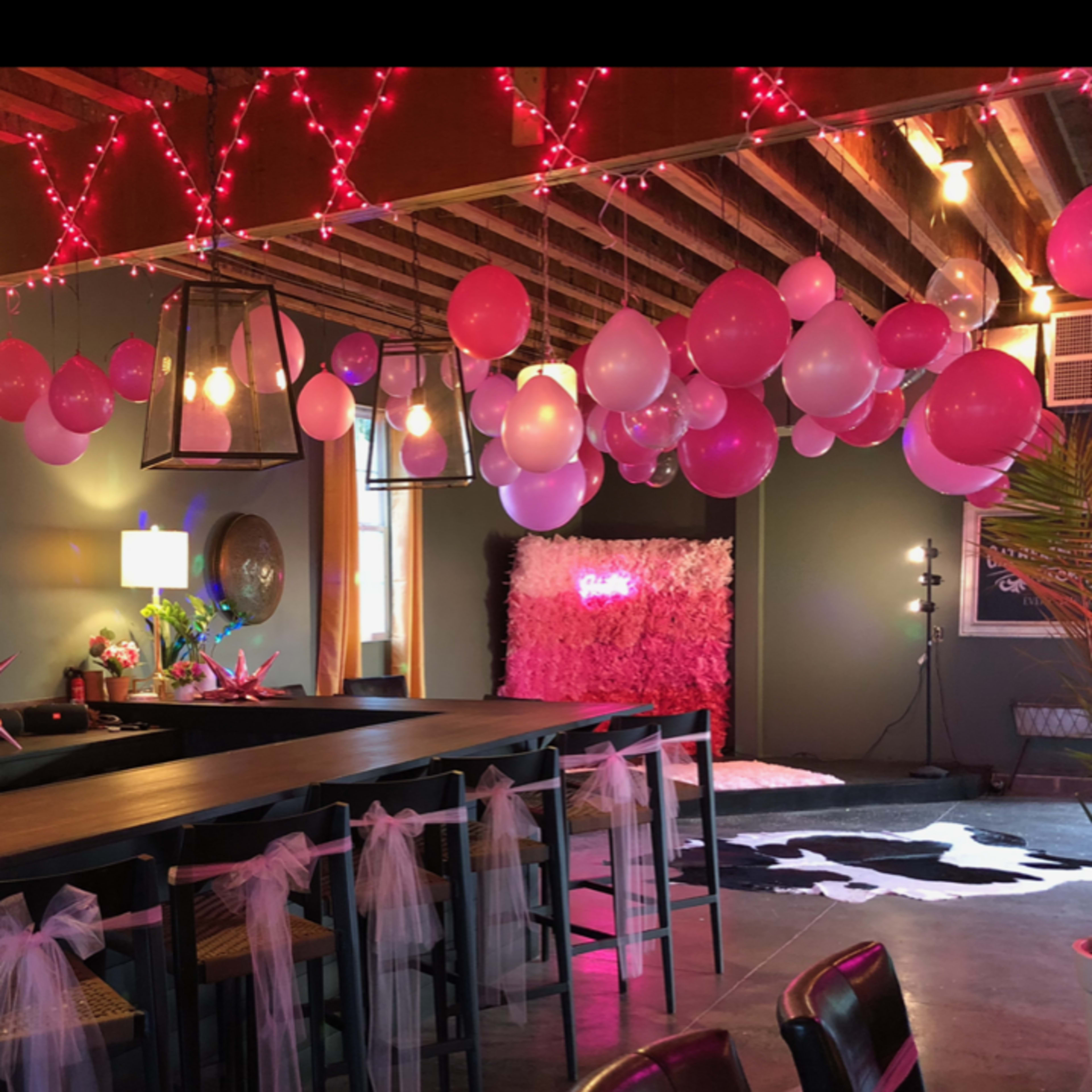 A decorated event space featuring pink balloons and fabric drapes hanging from the ceiling, with a long table adorned with sashes.