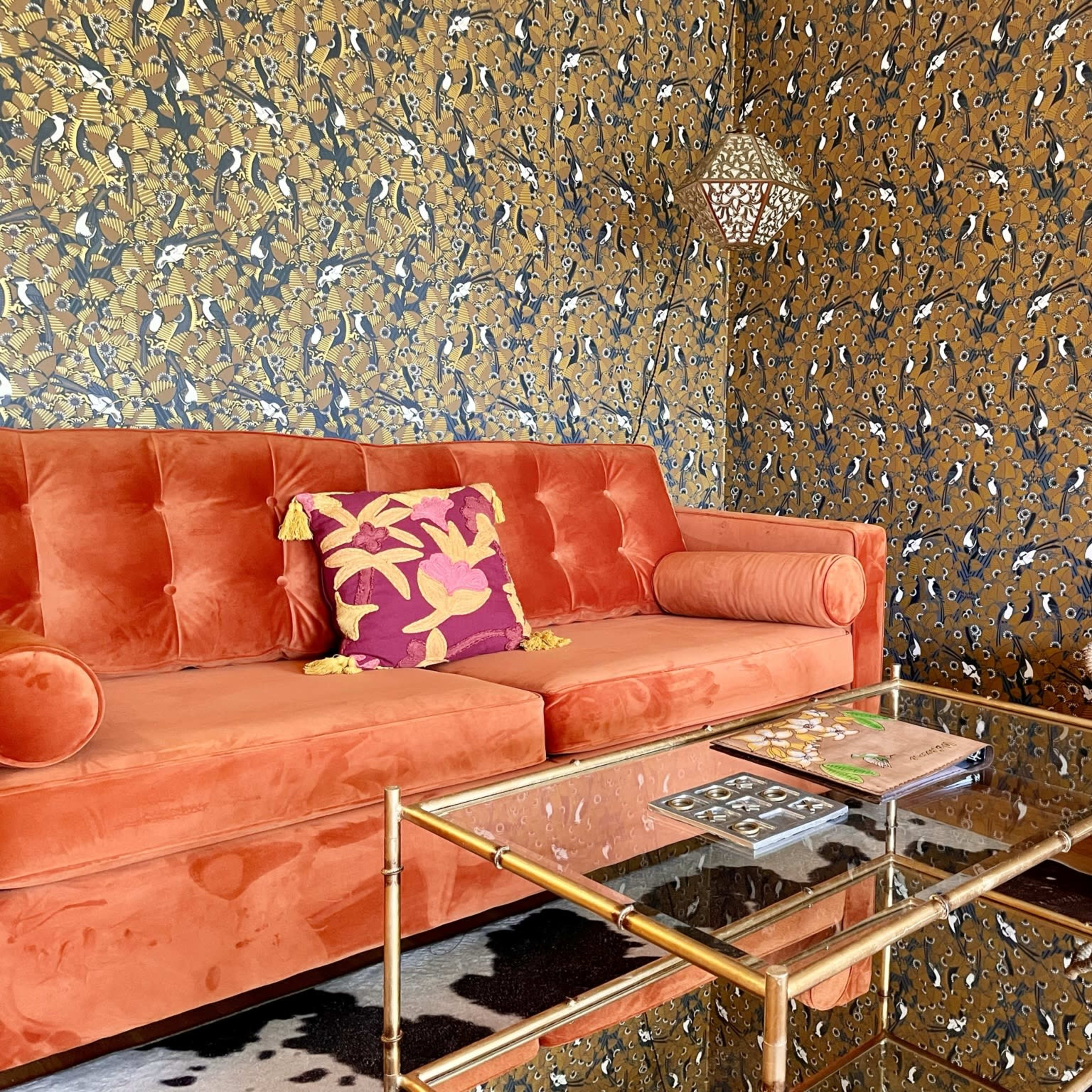 The scene features an orange velvet sofa with a decorative pillow in front of a patterned gold and blue wallpaper, accompanied by a glass coffee table and a woven basket on a cowhide rug.