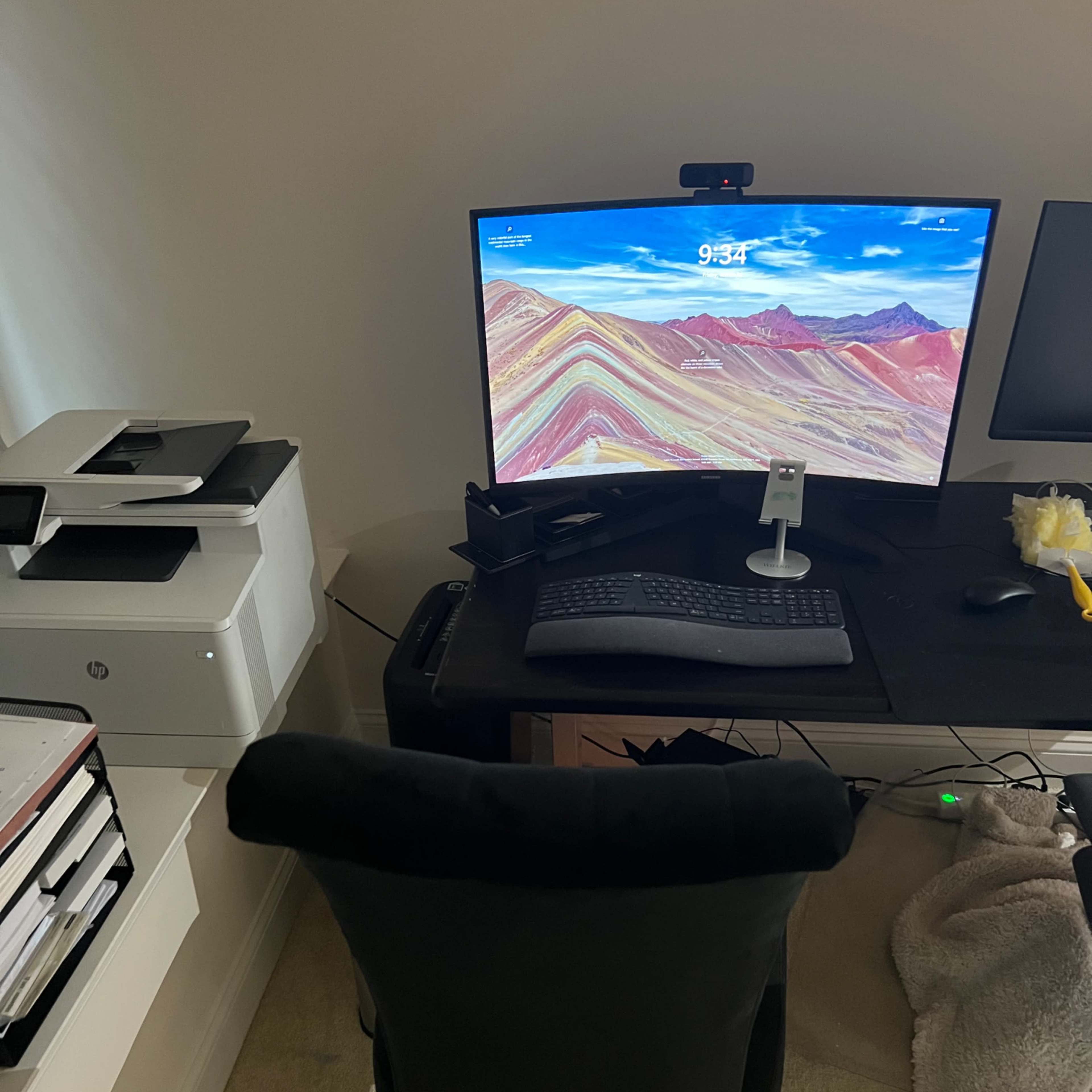 A desk with a computer setup featuring a large monitor, keyboard, and a black chair facing the screen, with a printer and files nearby.