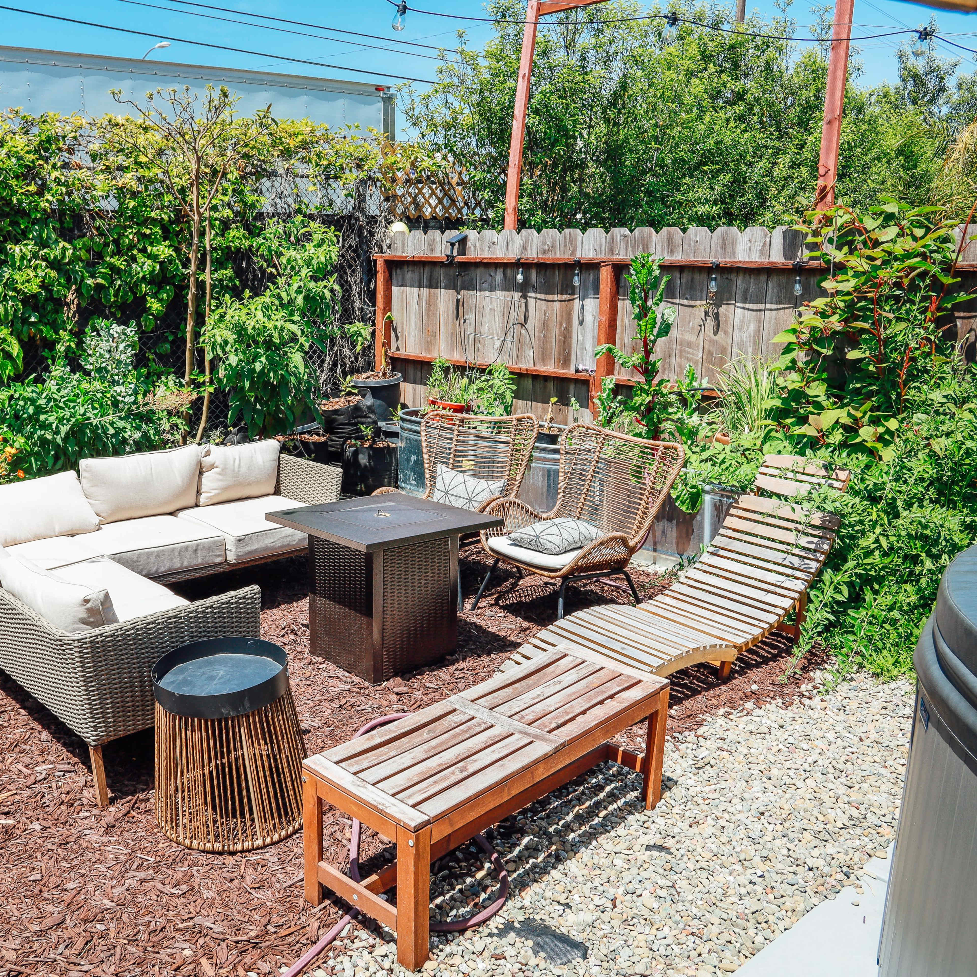 A landscaped backyard with a seating arrangement of wicker couches and chairs around a central table, surrounded by greenery and wood fencing.