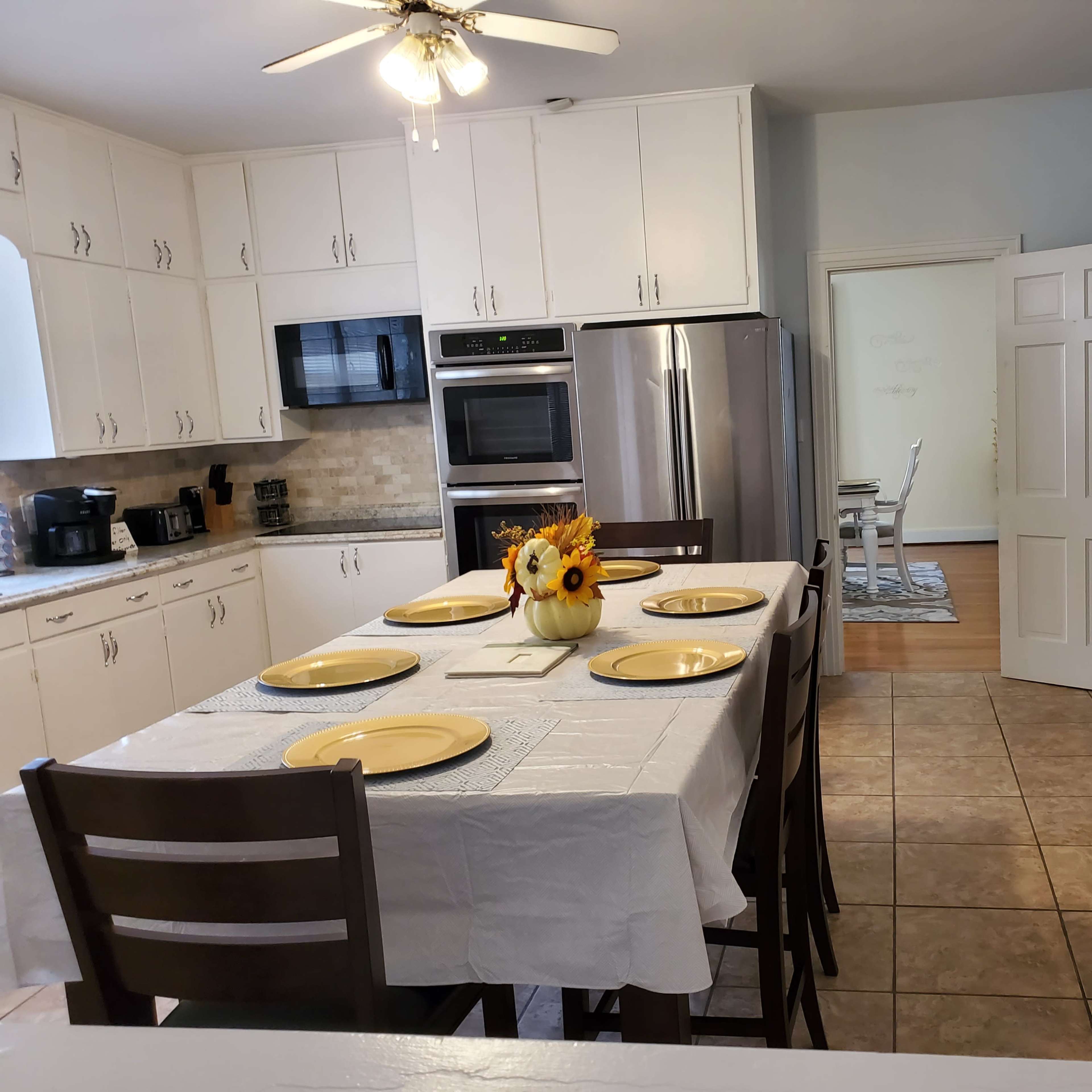 A kitchen features a table set for four with yellow plates, bright flowers in the center, and white cabinetry lining the walls.