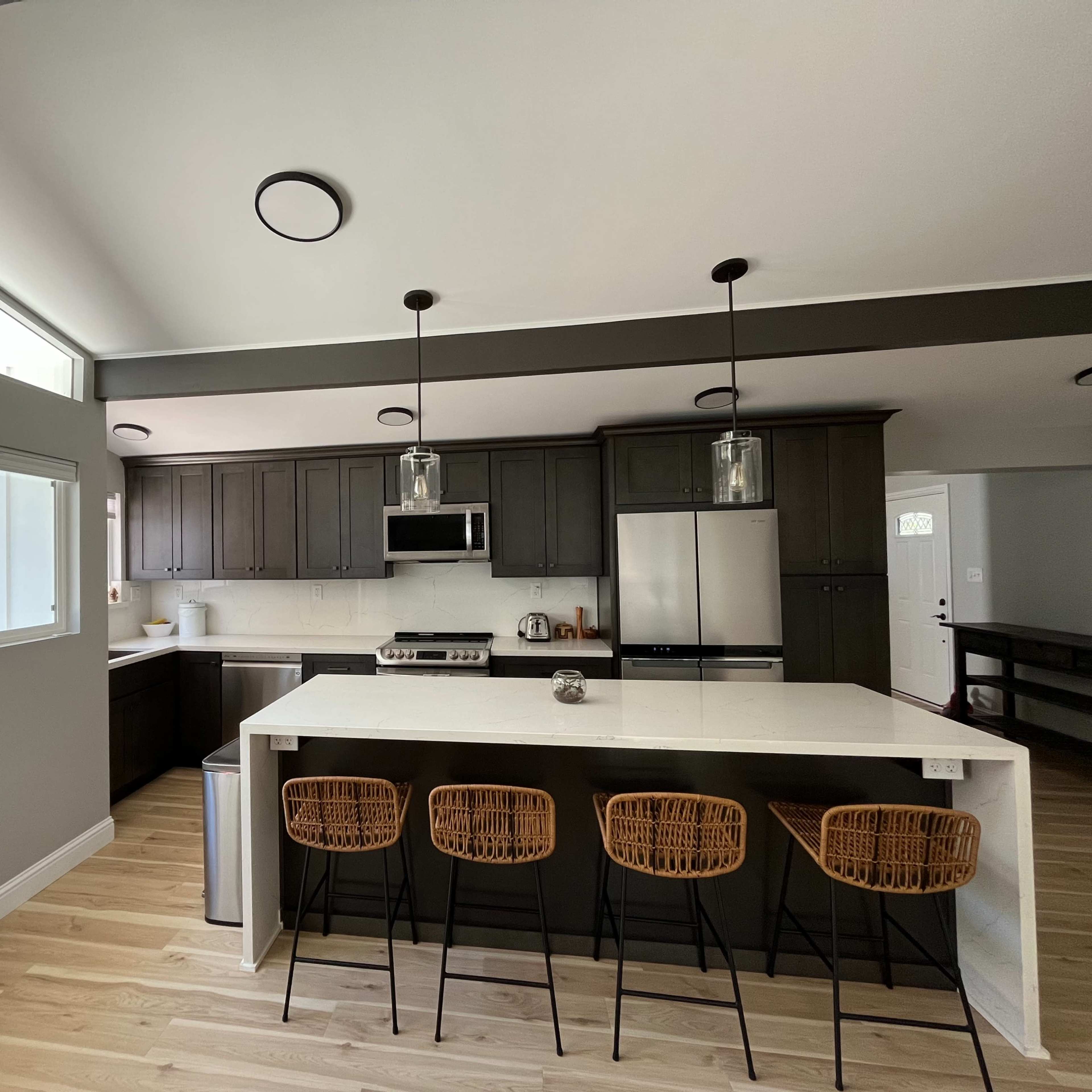 A modern kitchen featuring dark cabinets, stainless steel appliances, and a white island with four rattan barstools.