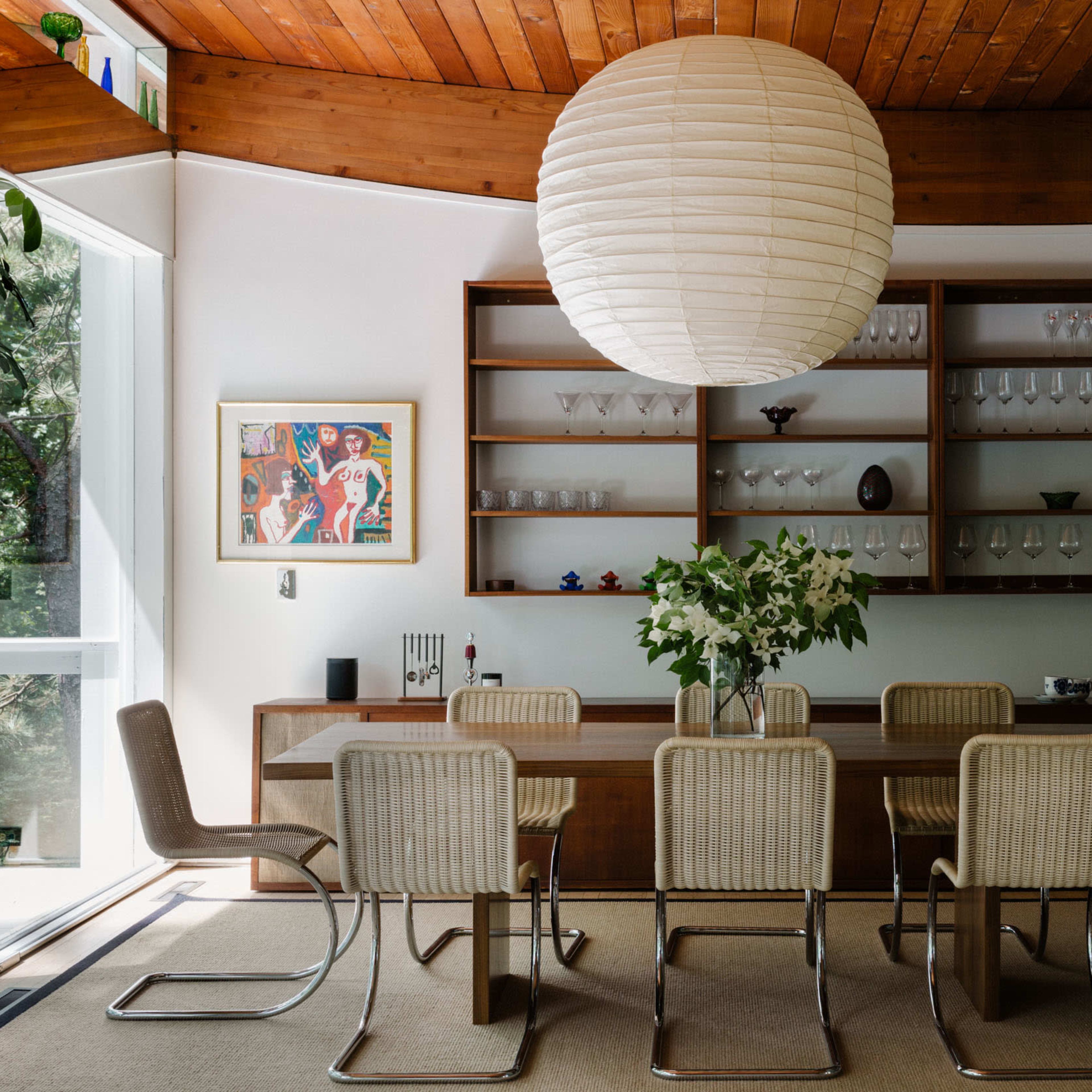A modern dining room features a large wooden table surrounded by six metal-framed chairs, illuminated by a round paper pendant light, with glassware displayed on shelves above.